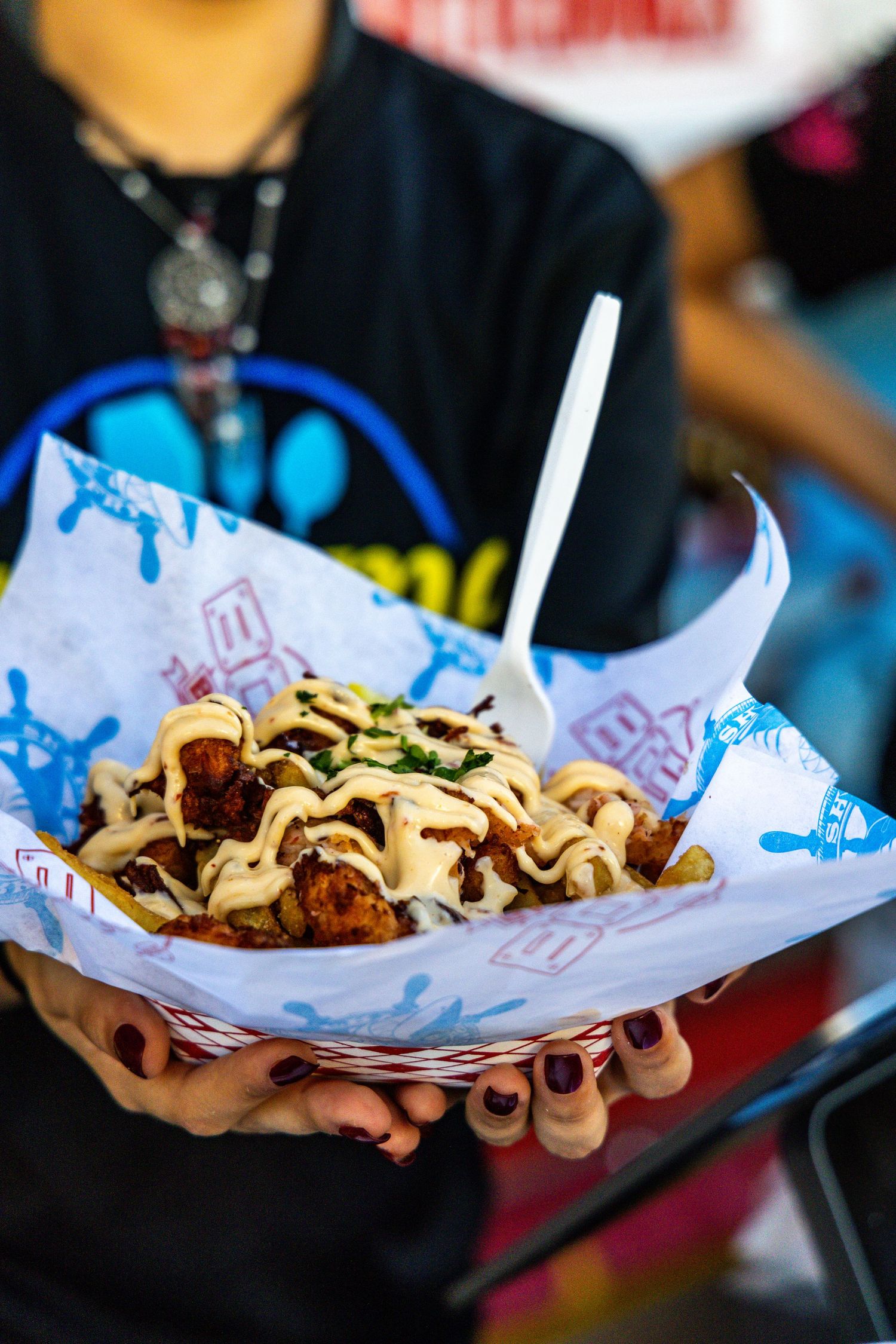 Hands holding a basket of loaded fries topped with creamy sauce and herbs at an outdoor food fair.