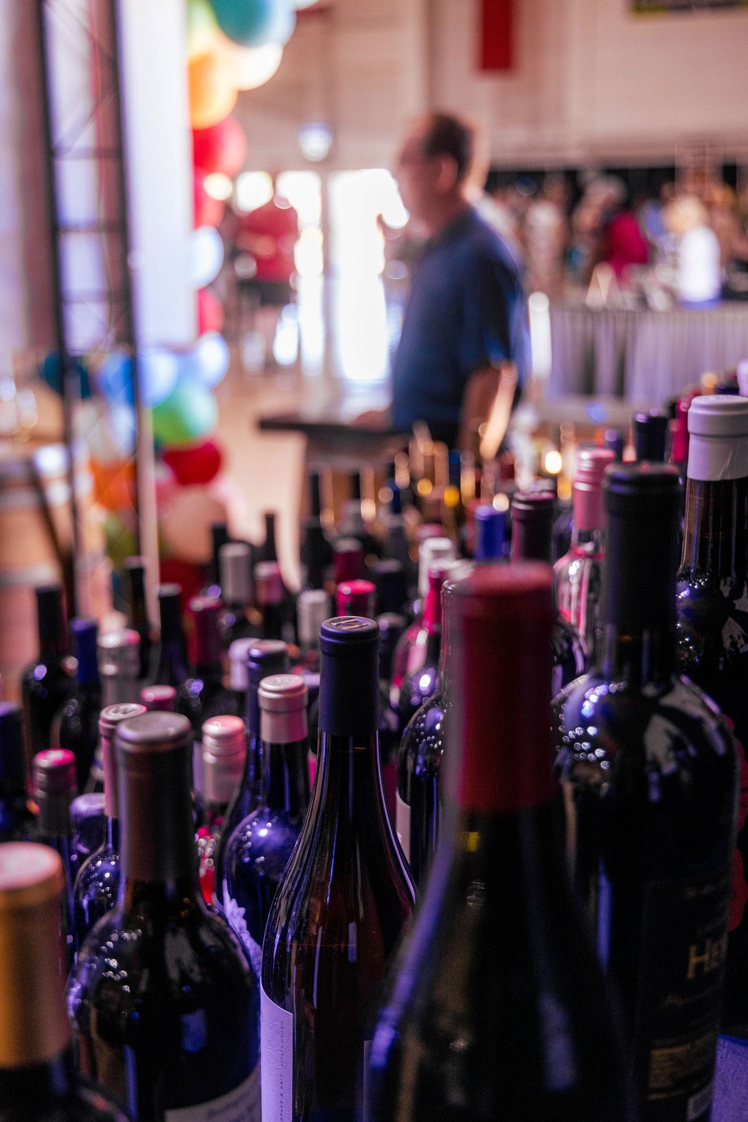 Rows of wine bottles in foreground with blurred man and colorful balloons in background at a wine tasting event.