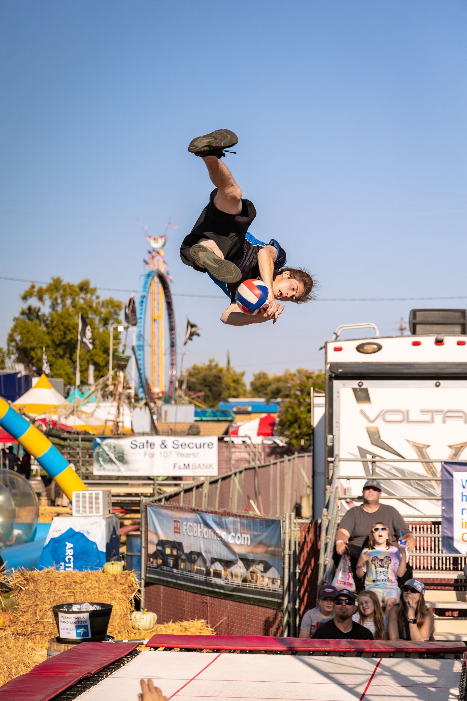 Acrobat performing a backflip with a ball at an outdoor county fair stunt show.