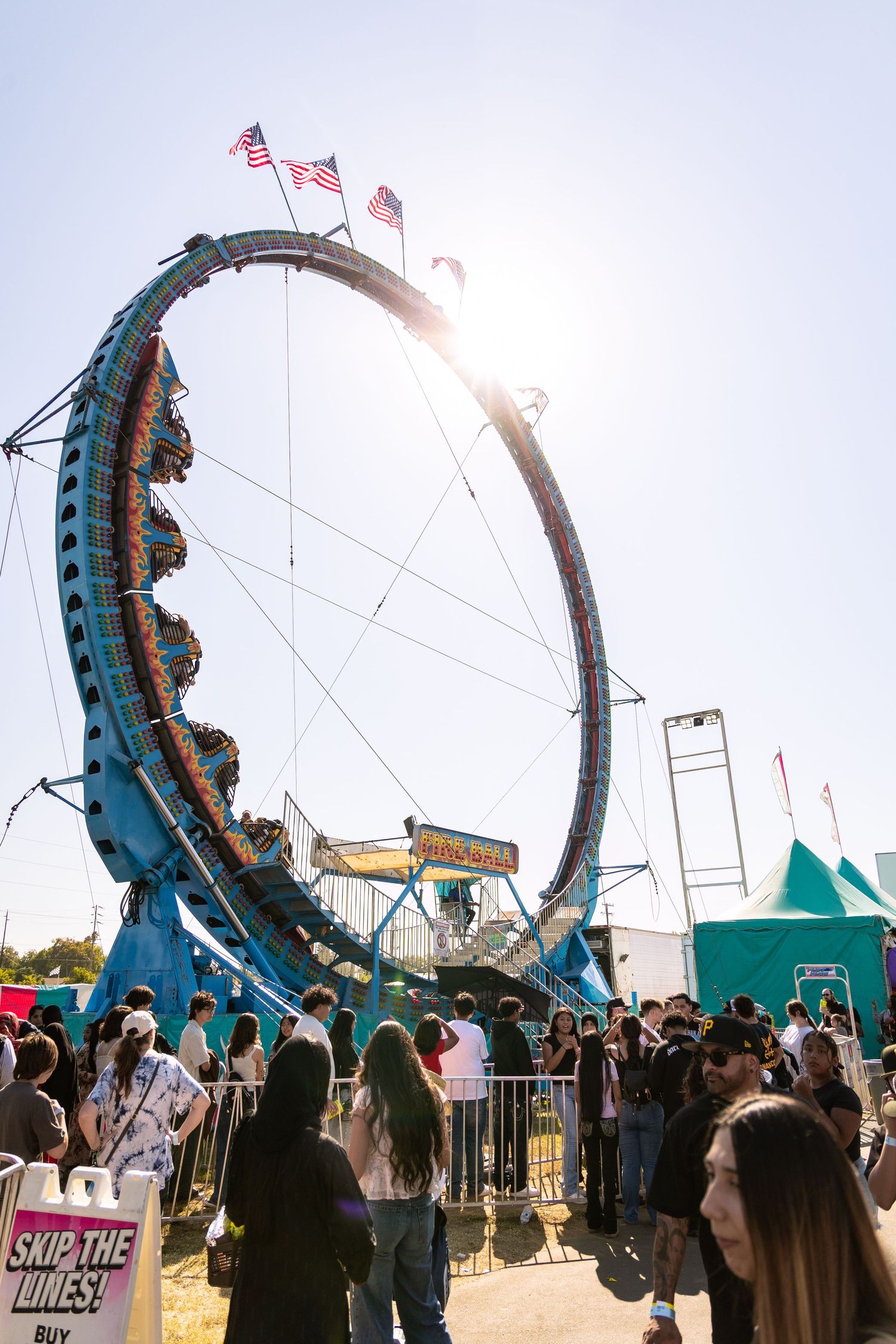 Crowd gathers around a large loop carnival ride at an outdoor fair on a sunny day.