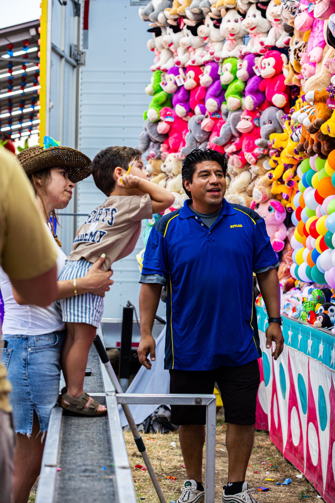 Carnival game booth worker stands near colorful stuffed animal prizes as family with child plays fair game.