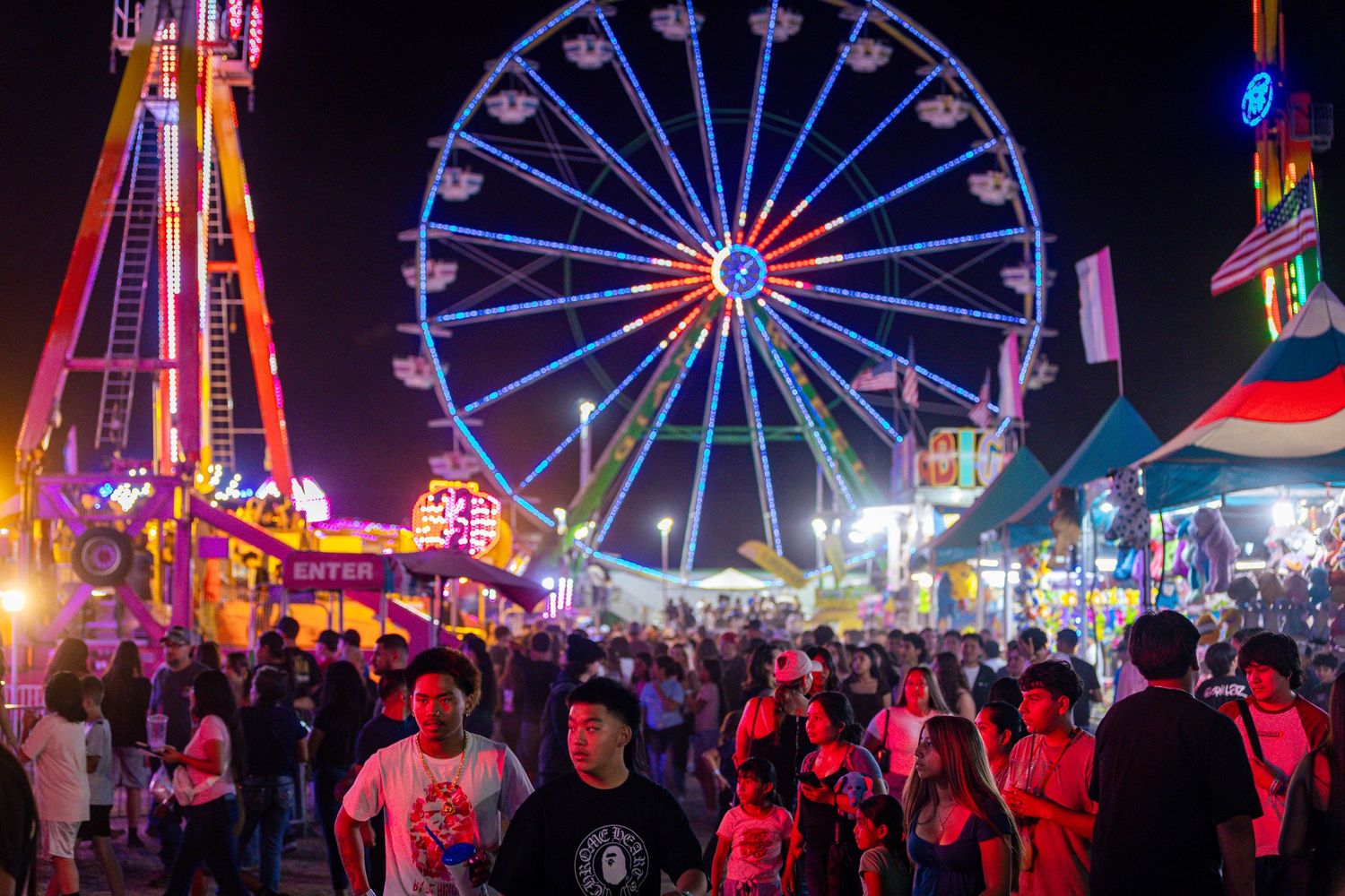 Large crowd enjoys a county fair at night with a glowing Ferris wheel and bright lights.