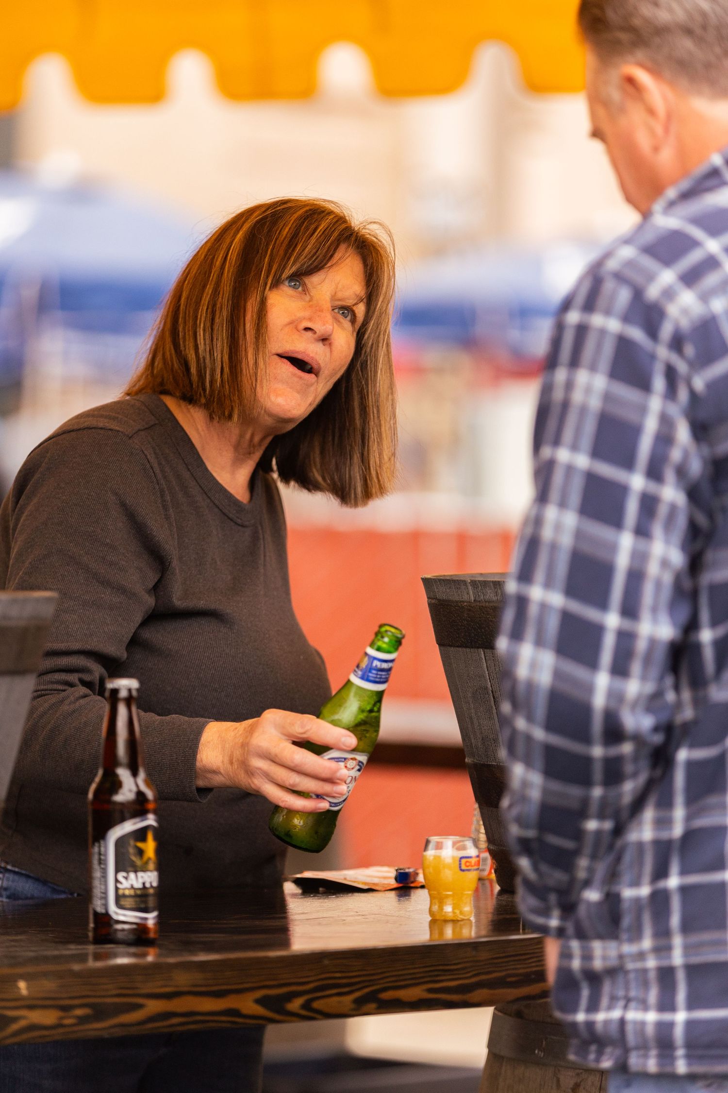 Woman holding a green beer bottle chatting with a man at an outdoor bar patio.