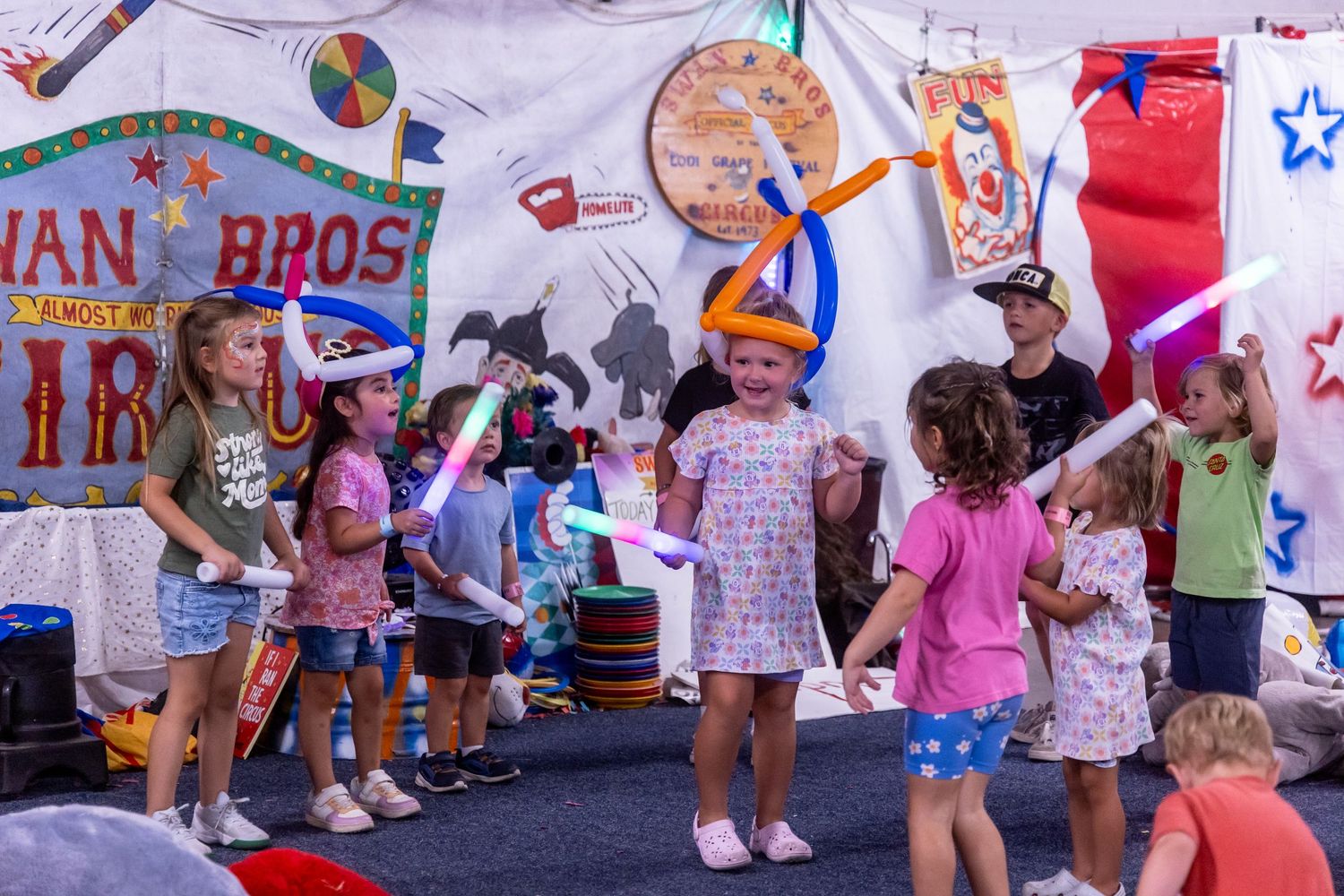 Children wearing balloon hats laugh and play with glow sticks at a lively circus tent performance.