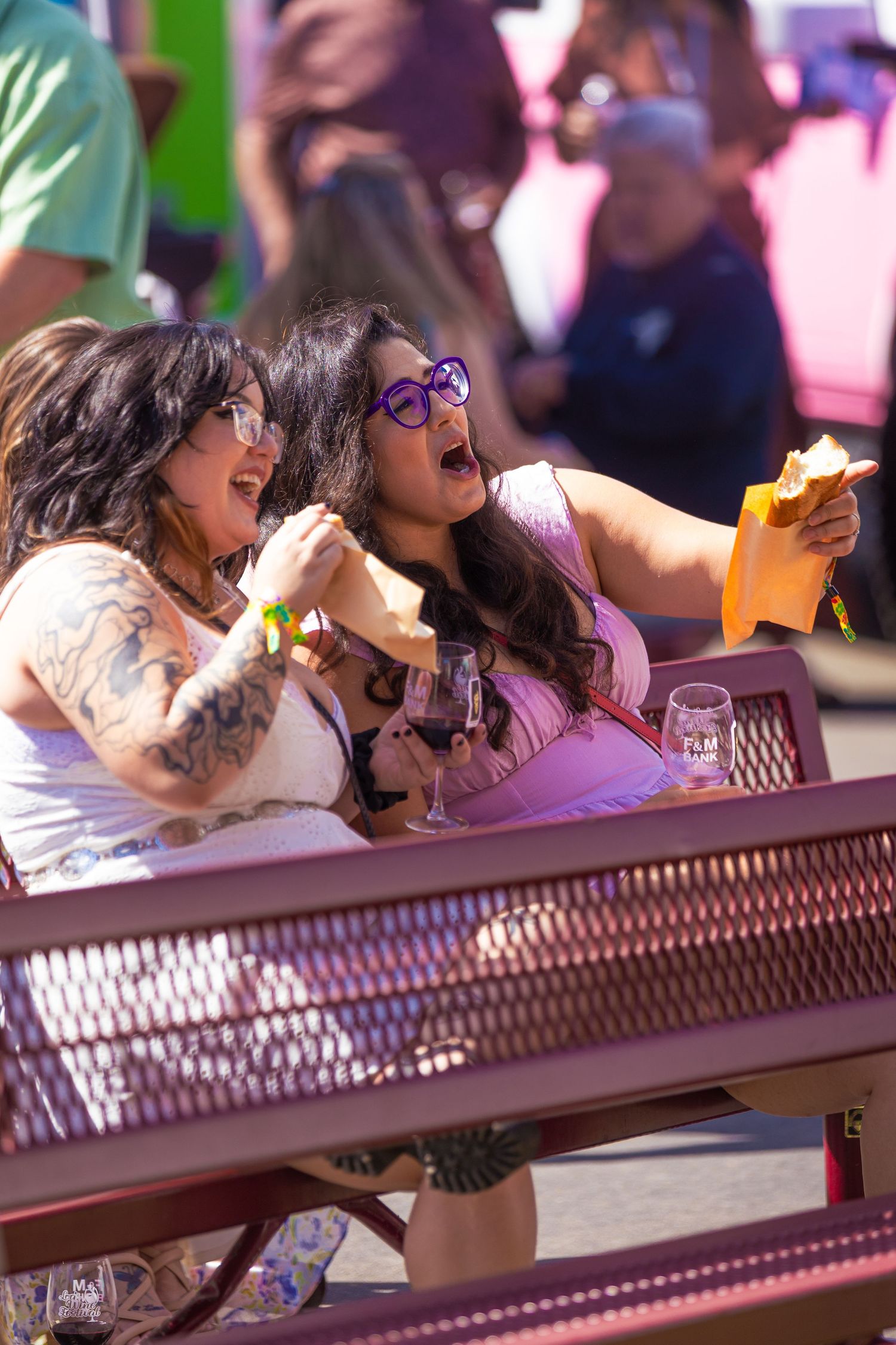 Two women laughing and enjoying food and wine while seated at a pink bench at an outdoor festival.