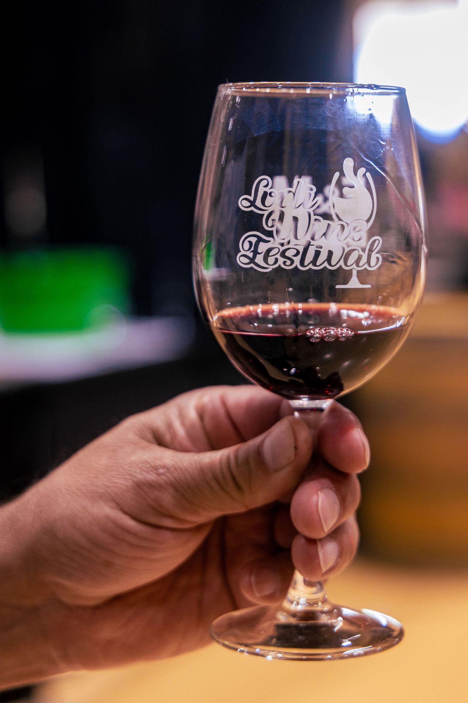 A hand holding a wine glass etched with 'Lake Wine Festival' logo, filled with red wine at a festival event.