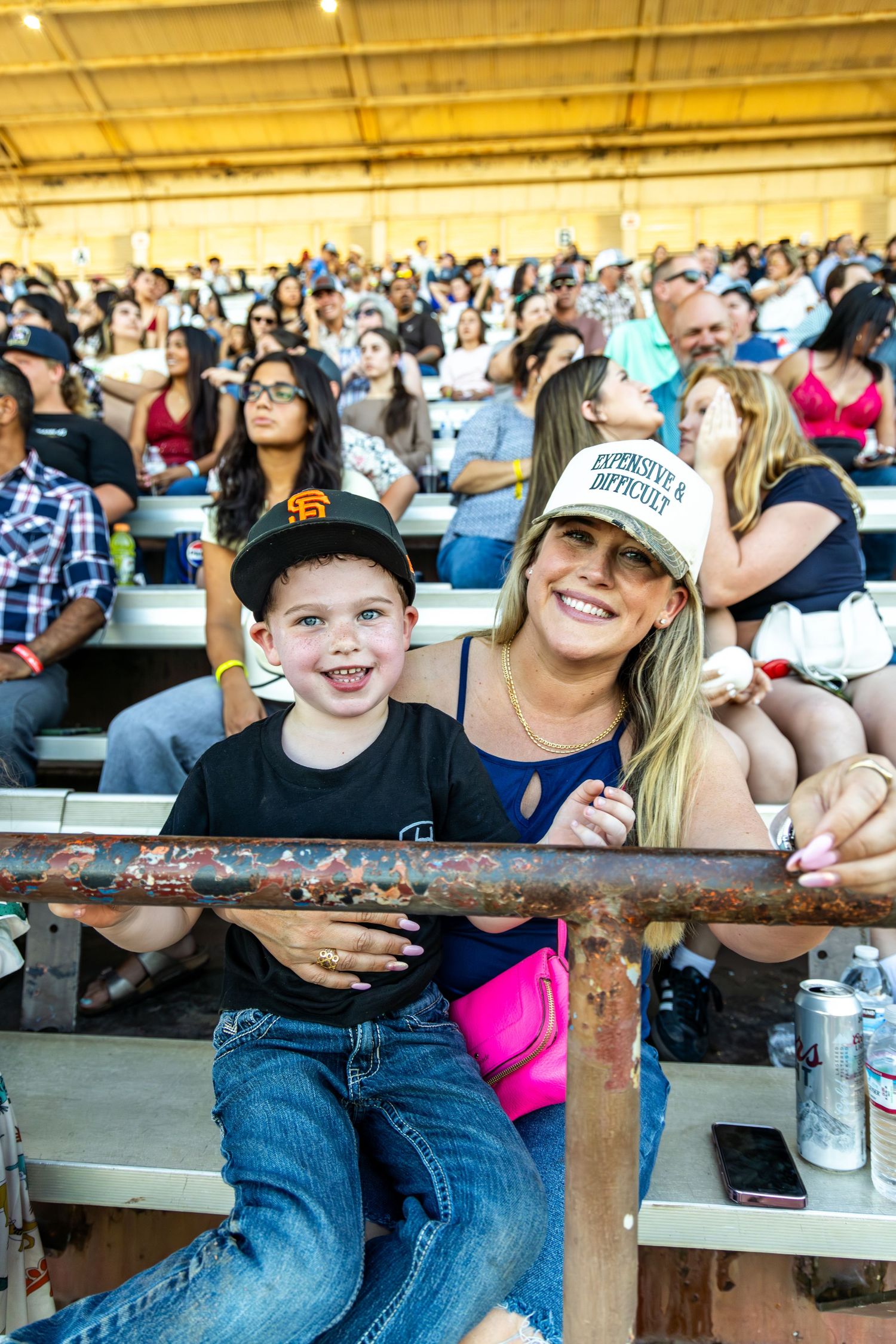 A smiling woman and young boy pose together in stadium bleachers during a crowded outdoor event.