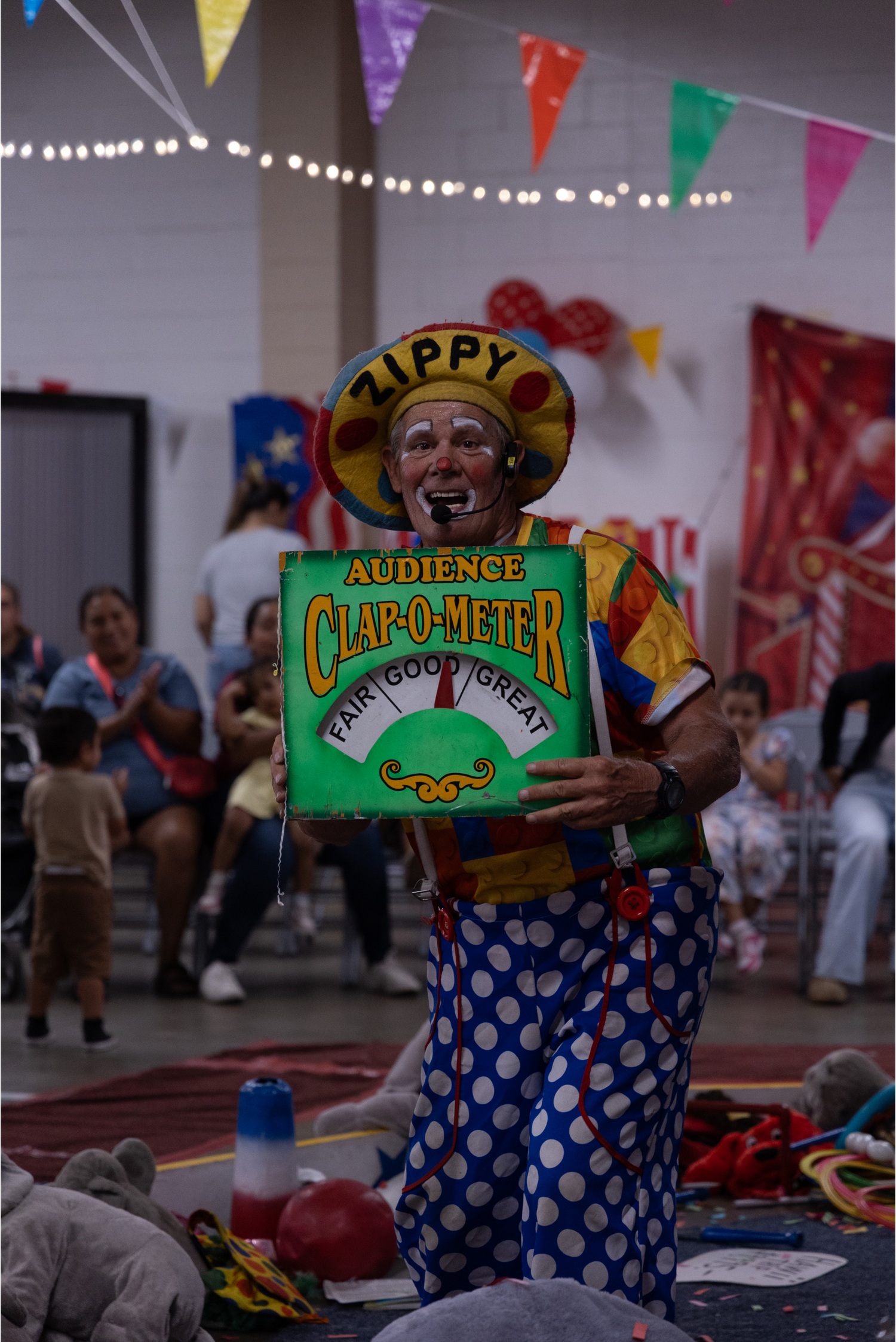 Clown named Zippy holds an Audience Clap-O-Meter sign at a colorful indoor circus performance.