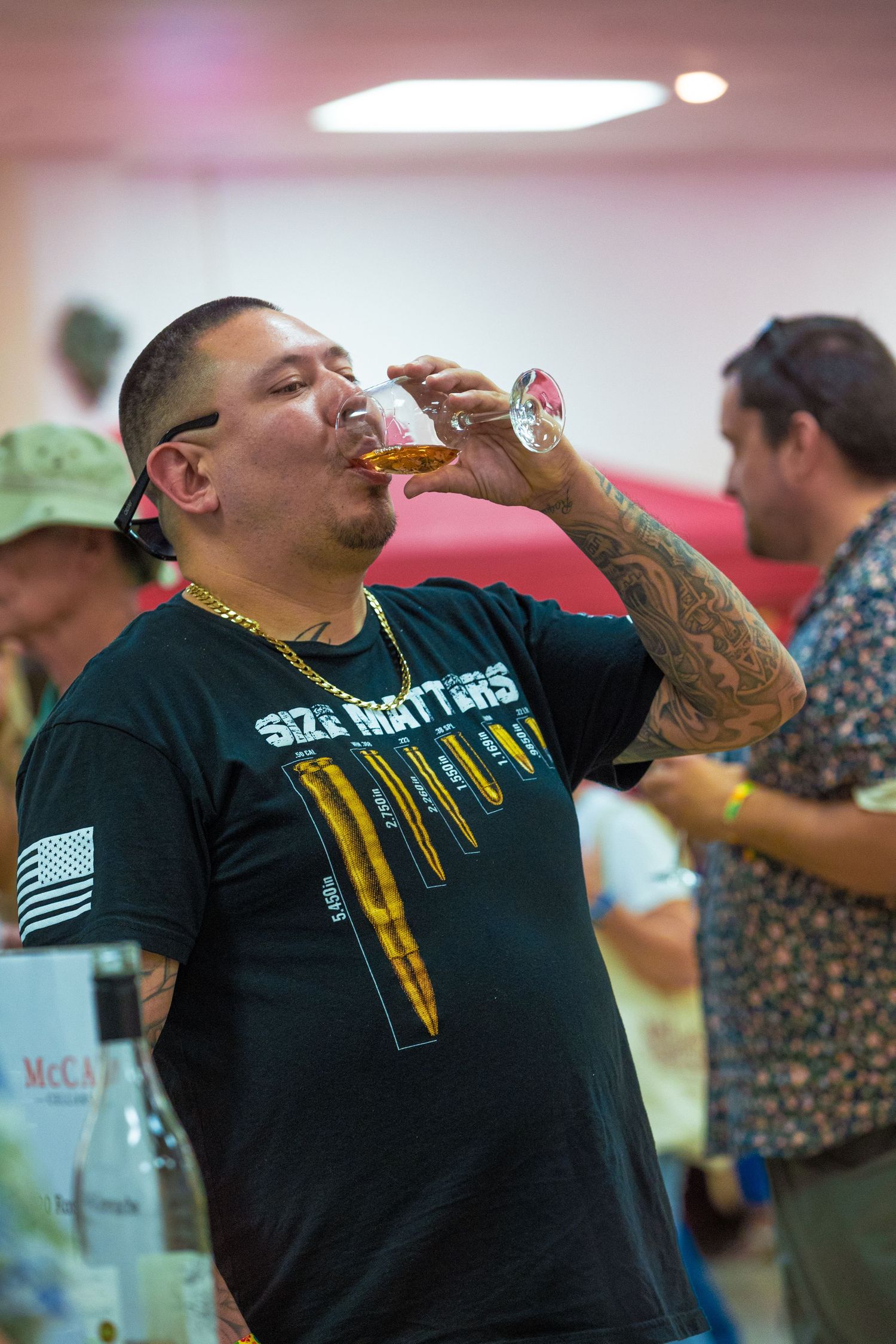 A tattooed man in a black t-shirt sampling whiskey at an indoor spirits tasting event.