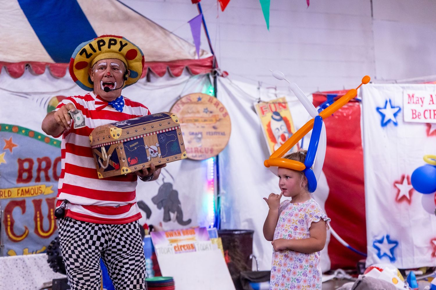 A clown named Zippy in a patriotic costume performs with a suitcase at a colorful circus tent show.