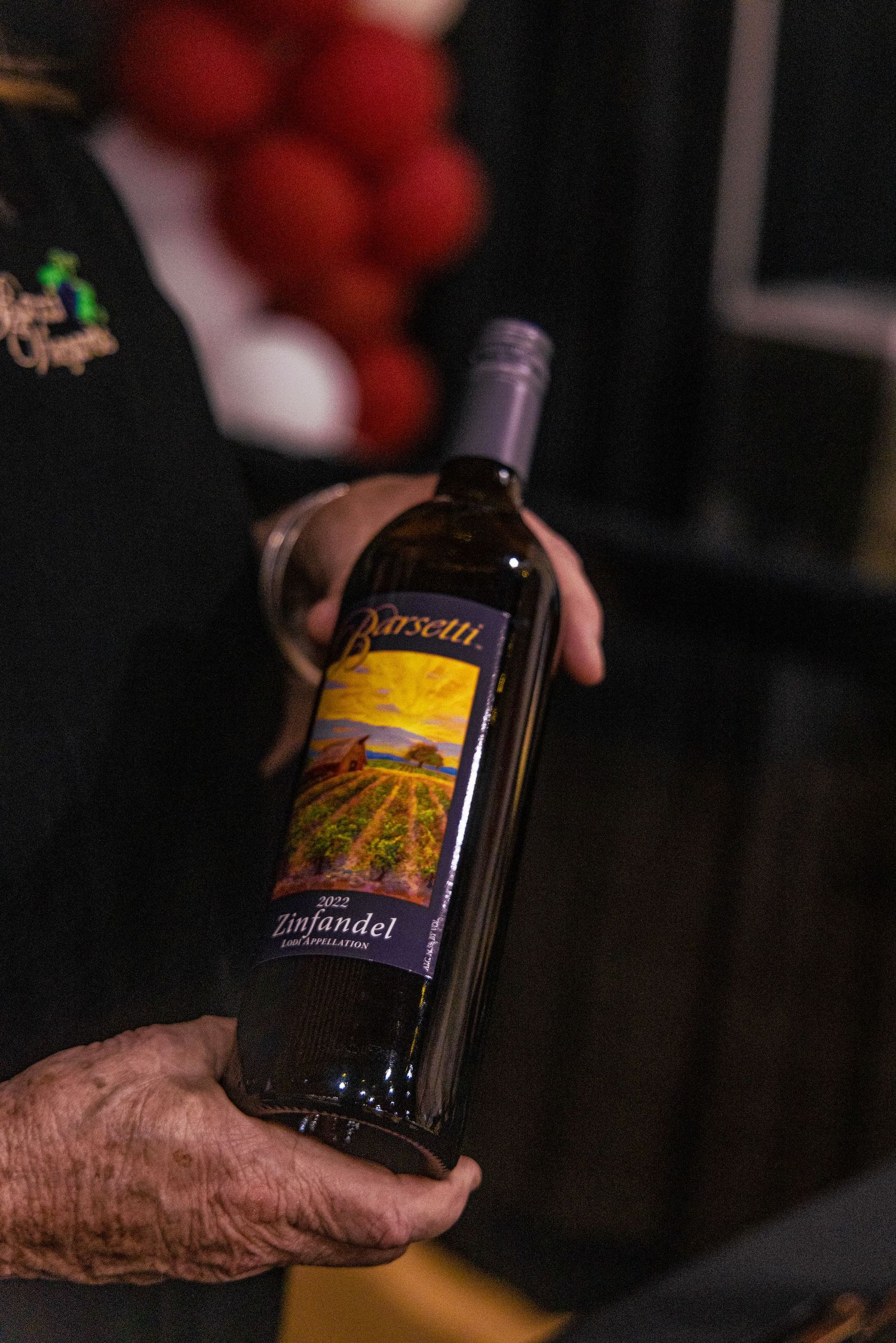 Hand holding a bottle of Barsetti 2022 Zinfandel wine with a colorful vineyard landscape label.
