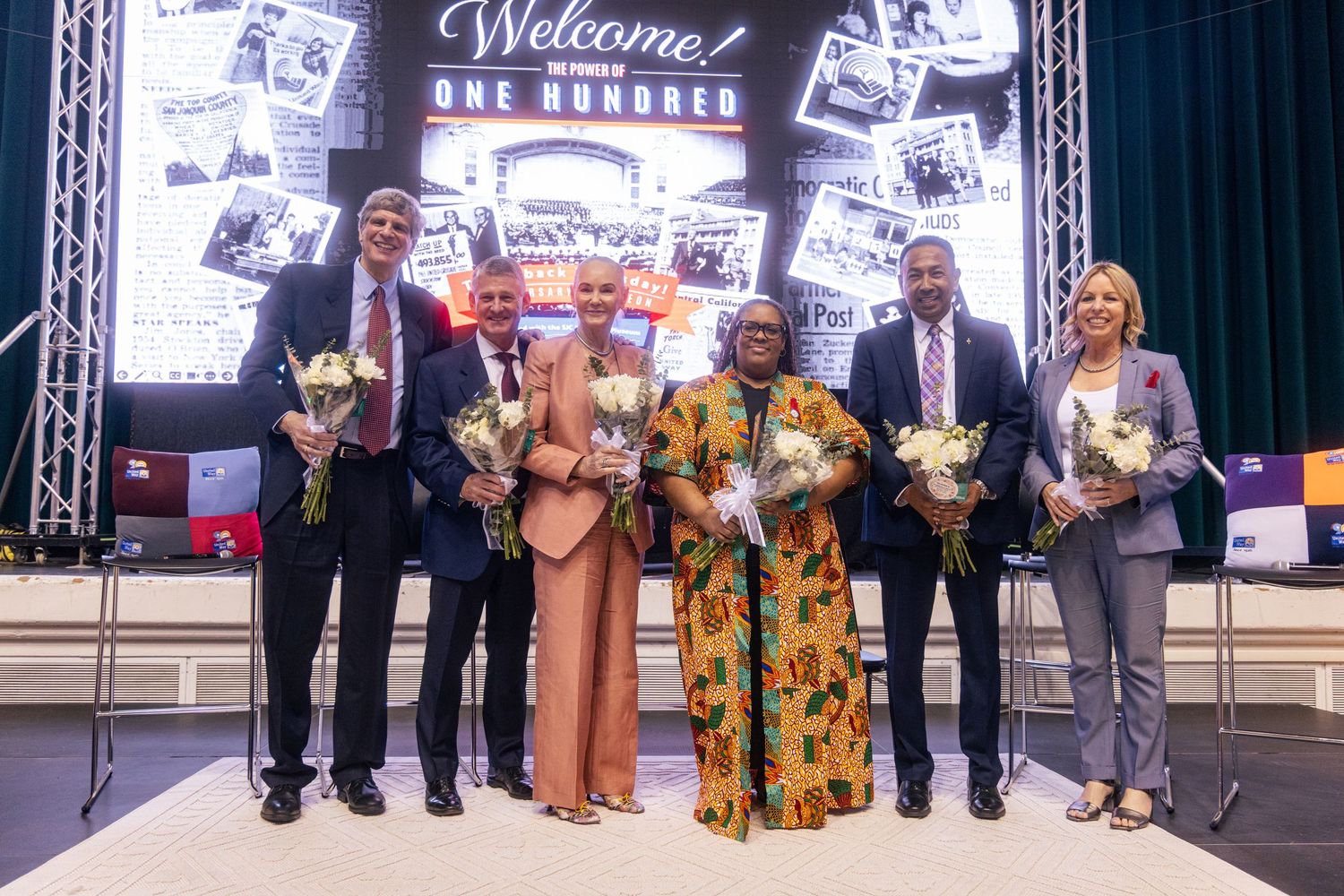 Six people holding flowers pose on stage at a Welcome One Hundred celebration event with a decorative backdrop.