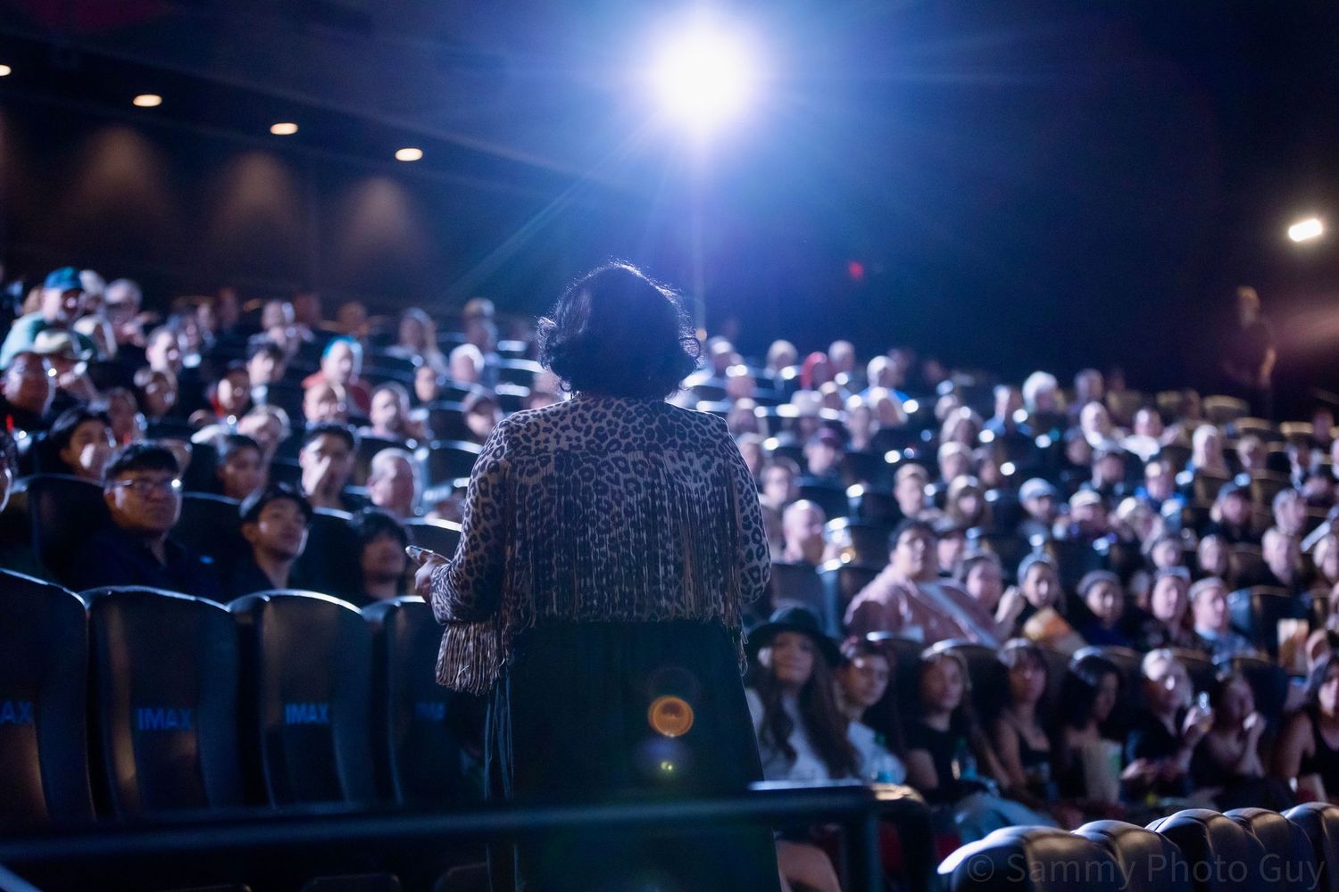 Person standing before a packed theater audience, viewed from behind, with bright stage lighting illuminating the crowd.