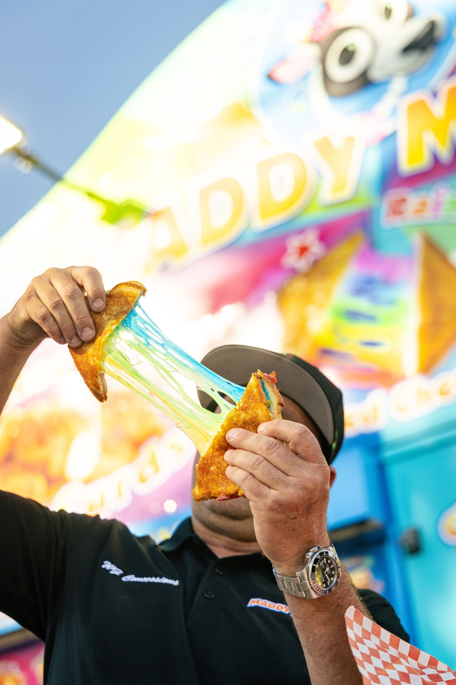 Man pulls apart a cheesy fried food item in front of a colorful fair food stand.