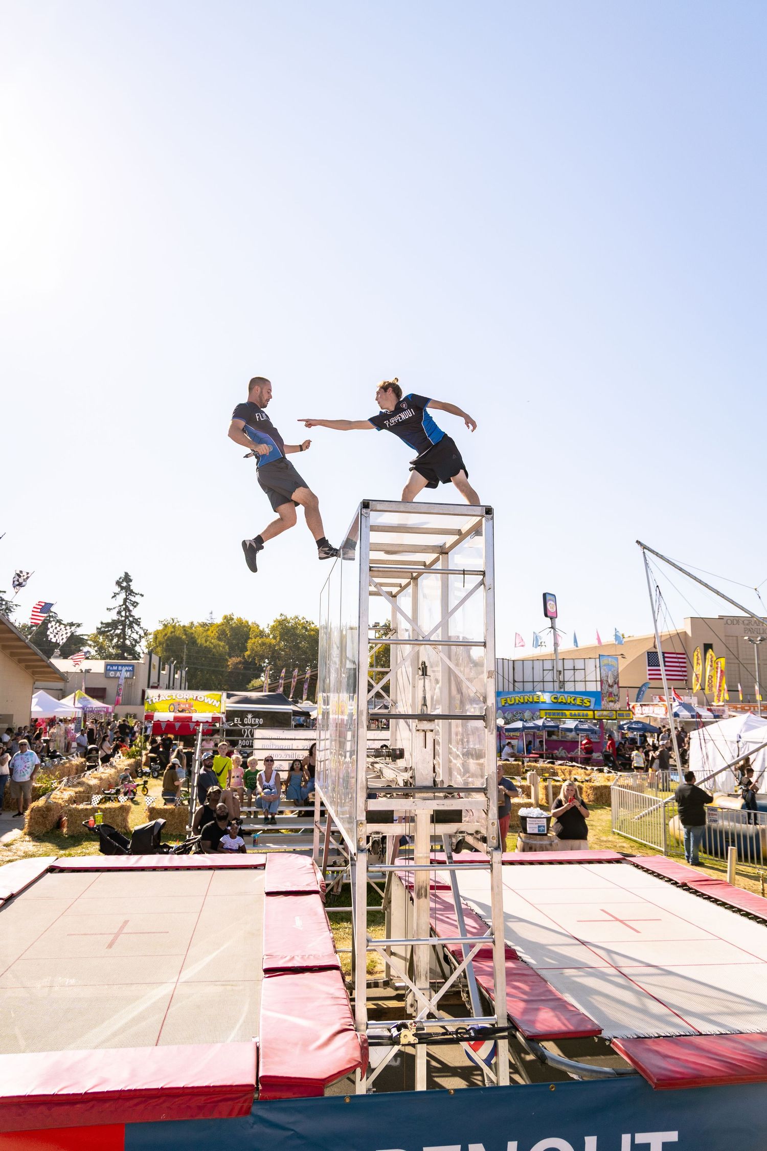 Two performers leap from a metal tower structure at an outdoor fair stunt show.