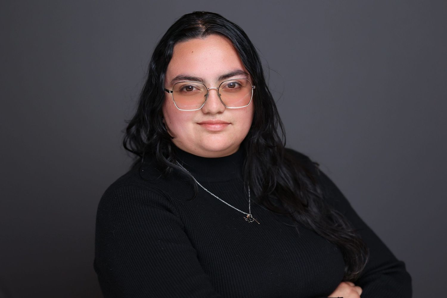 Young woman with long black hair and glasses wearing a black turtleneck, arms crossed, professional headshot.