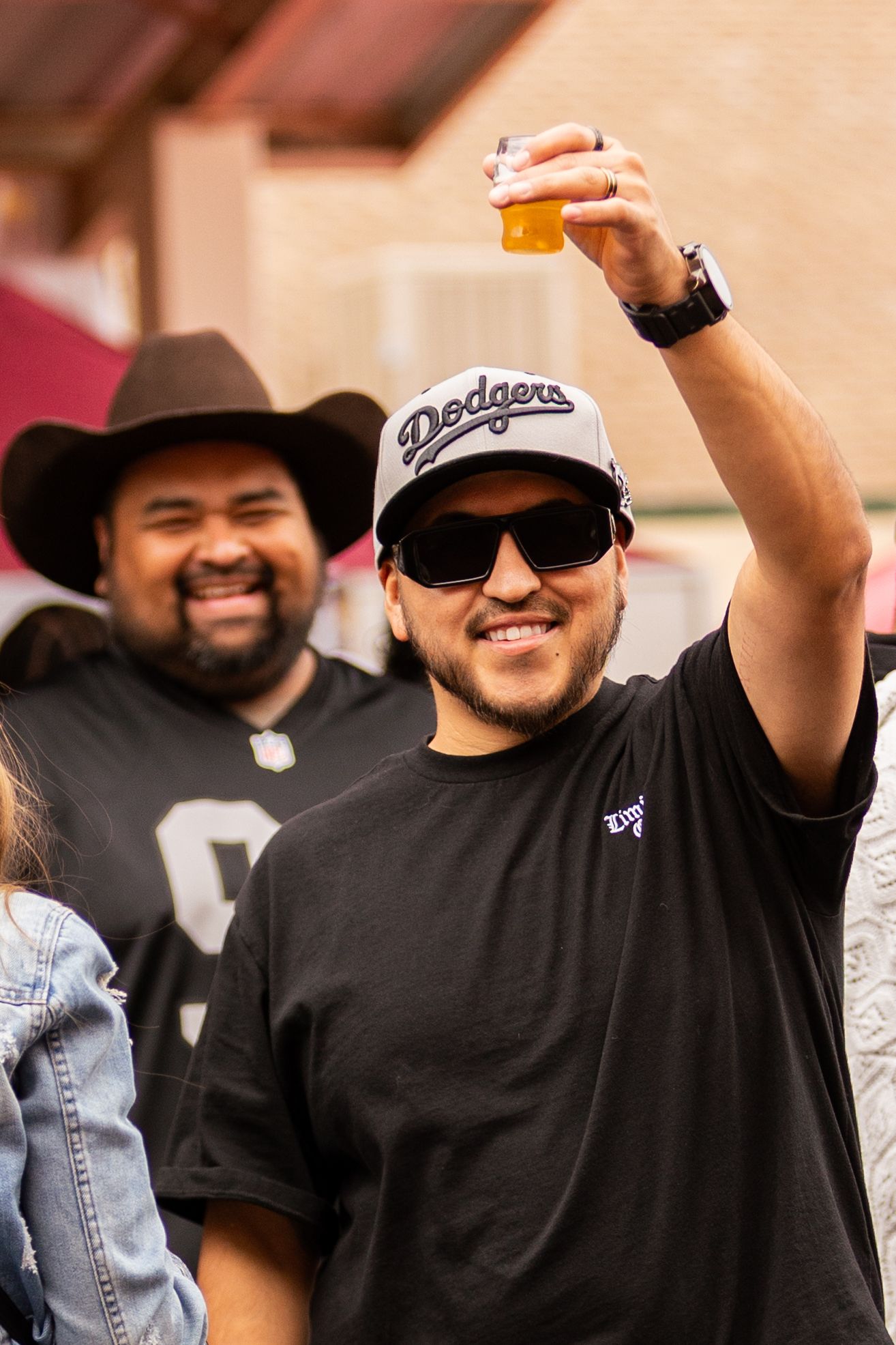 Man in Dodgers cap raises beer glass at outdoor gathering with friends wearing cowboy hat and Raiders jersey.