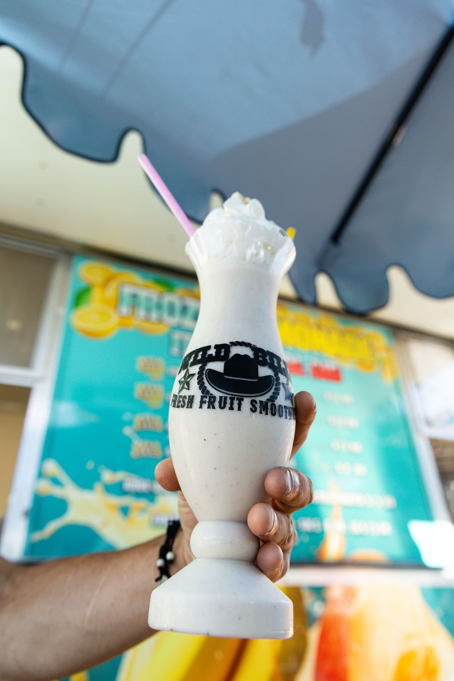 Hand holding a creamy milkshake in a tall glass with whipped cream and a pink straw at a fresh fruit smoothie stand.