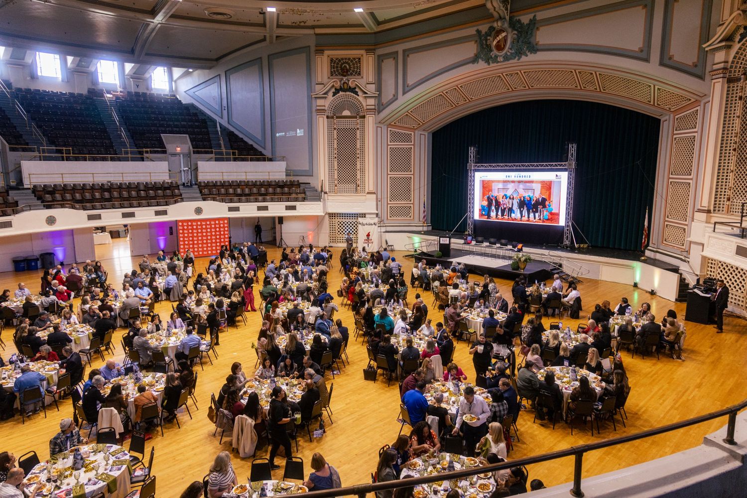 Elegant banquet hall with round tables of guests dining beneath a grand stage with presenters at a formal gala event.