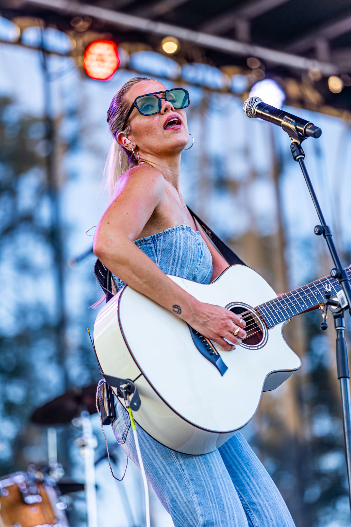Blonde female musician performing on stage wearing sunglasses and denim outfit while playing white acoustic guitar at outdoor concert.