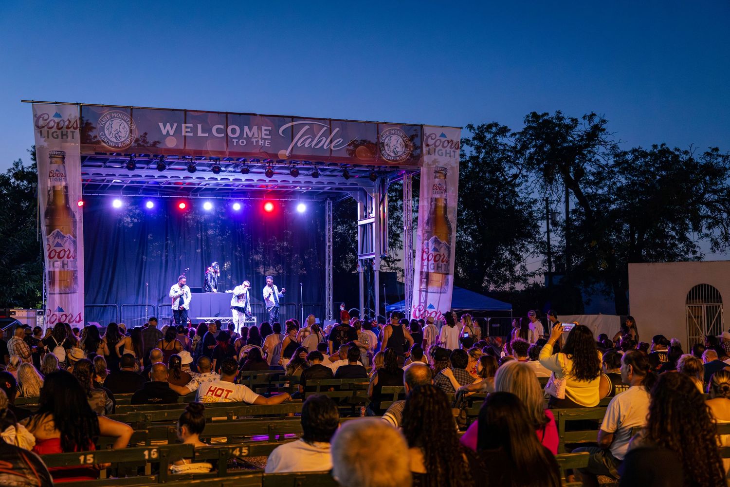 Outdoor concert stage with performers at dusk under a Welcome sign with seated audience at a festival.