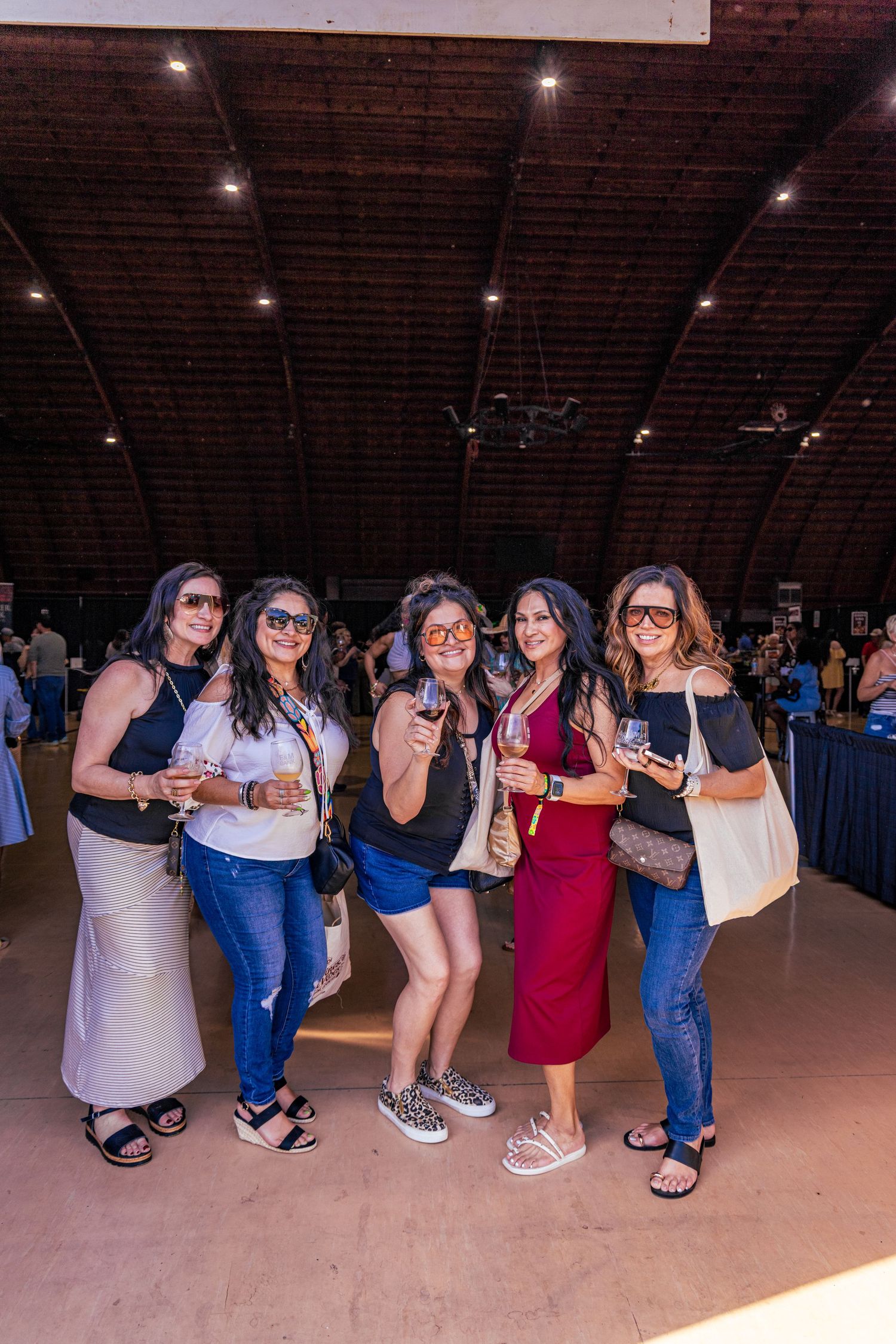 Group of five women smiling and holding drinks at an indoor wine tasting event in a large venue.
