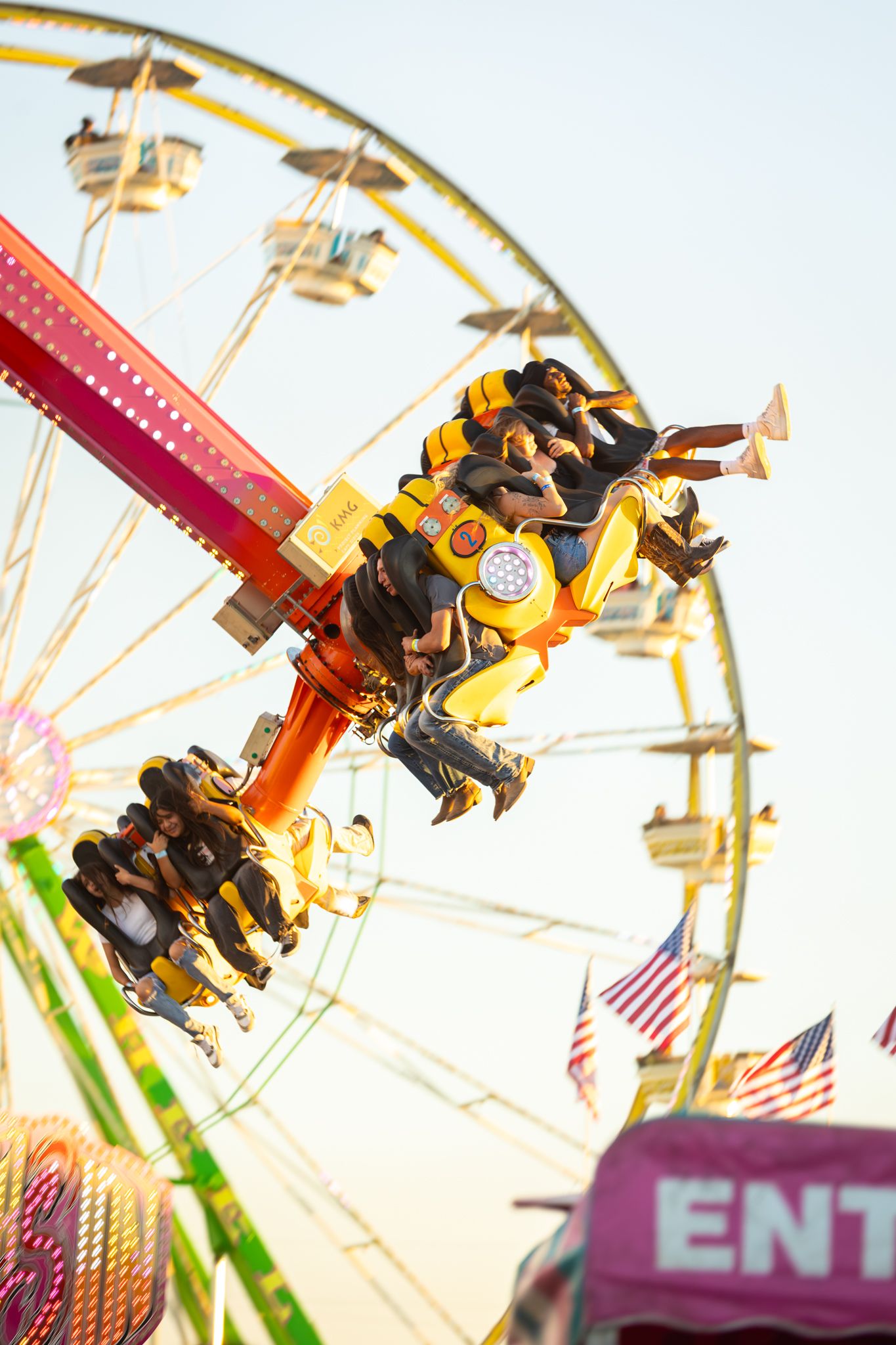 Thrilling carnival ride spinning riders upside down with a Ferris wheel and American flags in the background at a fairground.