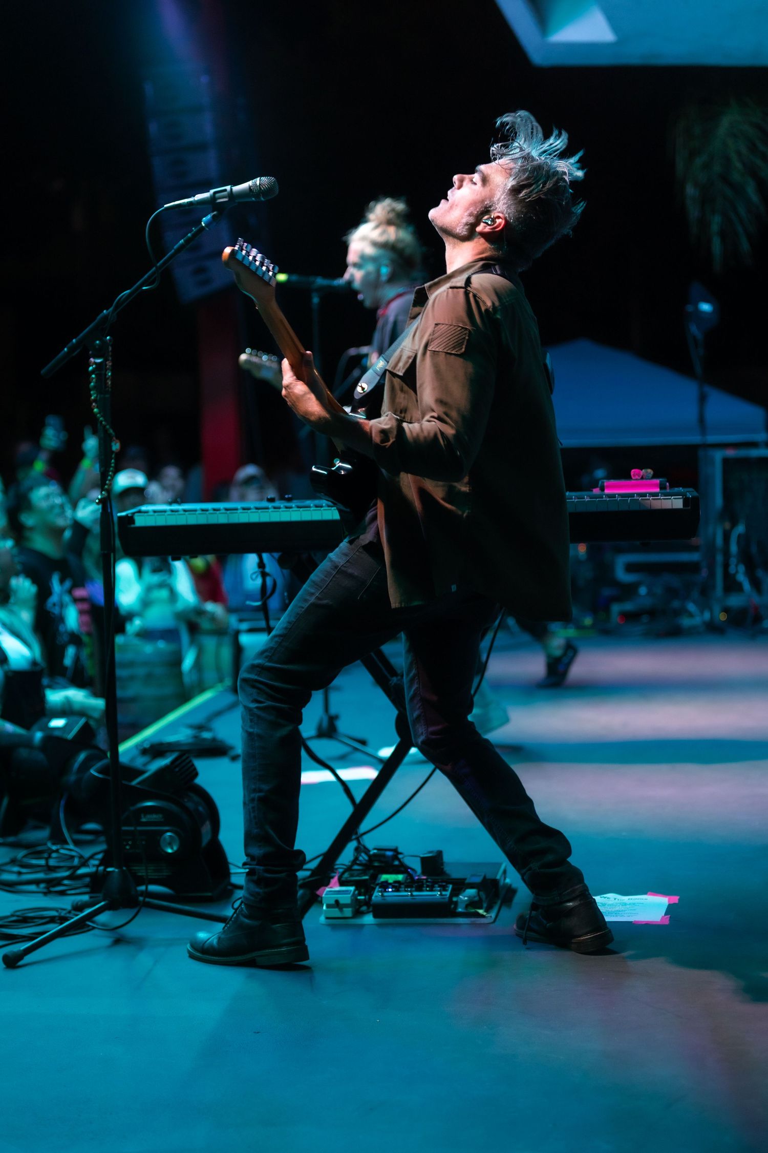 Electric guitarist leans back dramatically on stage with teal stage lighting and crowd in background.