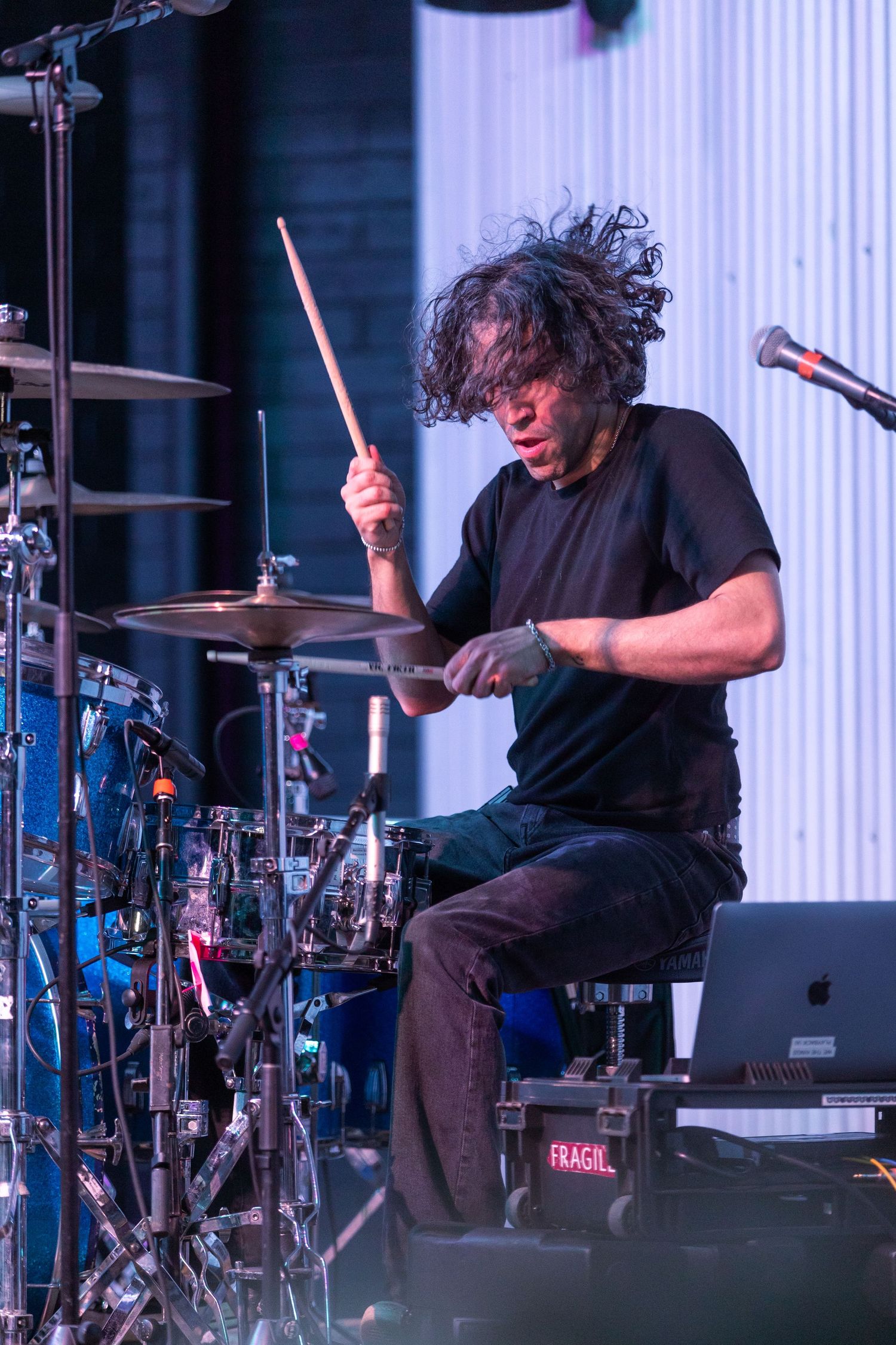Curly-haired drummer plays intensely on a full drum kit on stage under bright stage lighting.