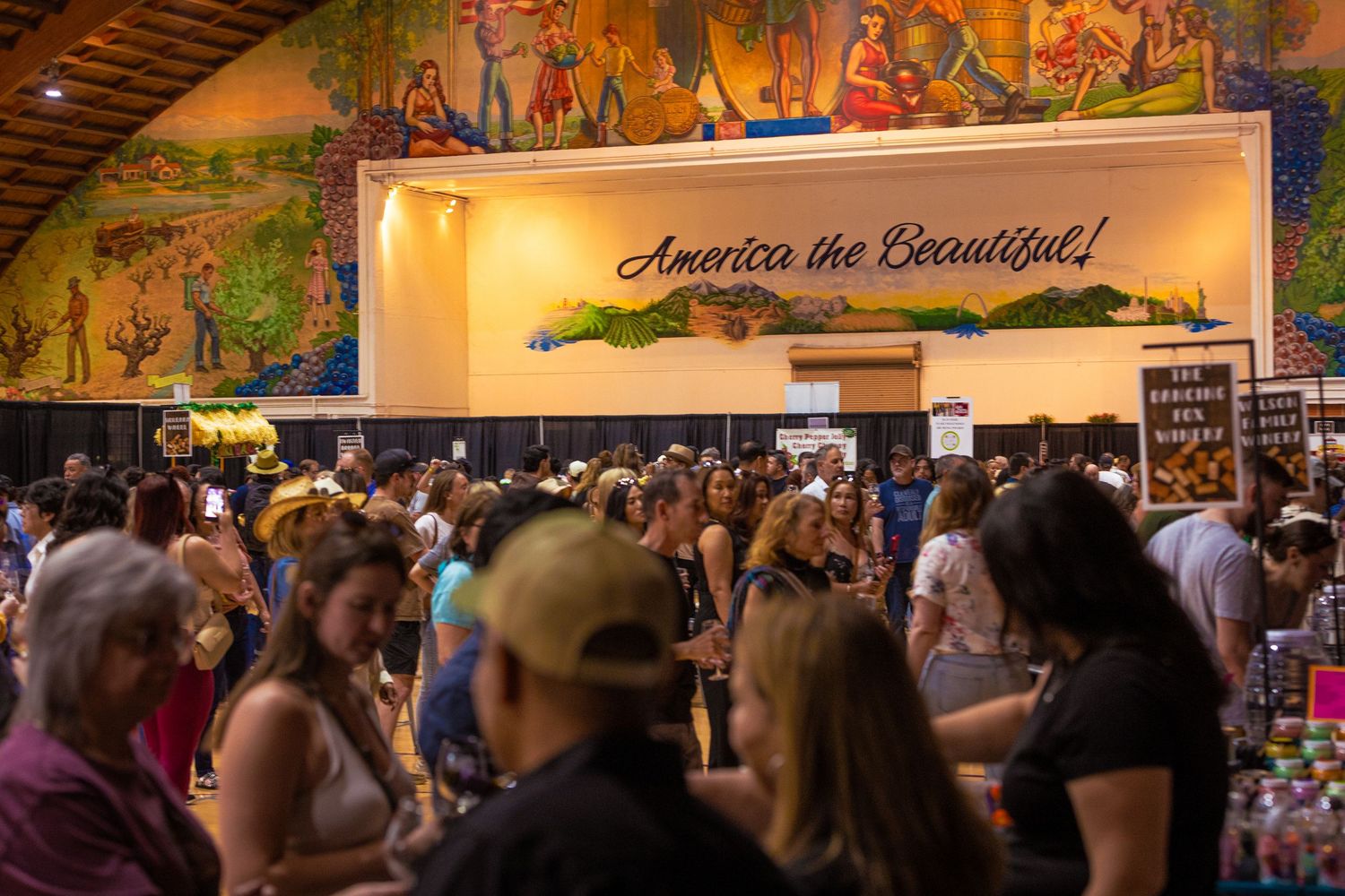 A large crowd of people browsing vendor booths inside a hall with an America the Beautiful mural.