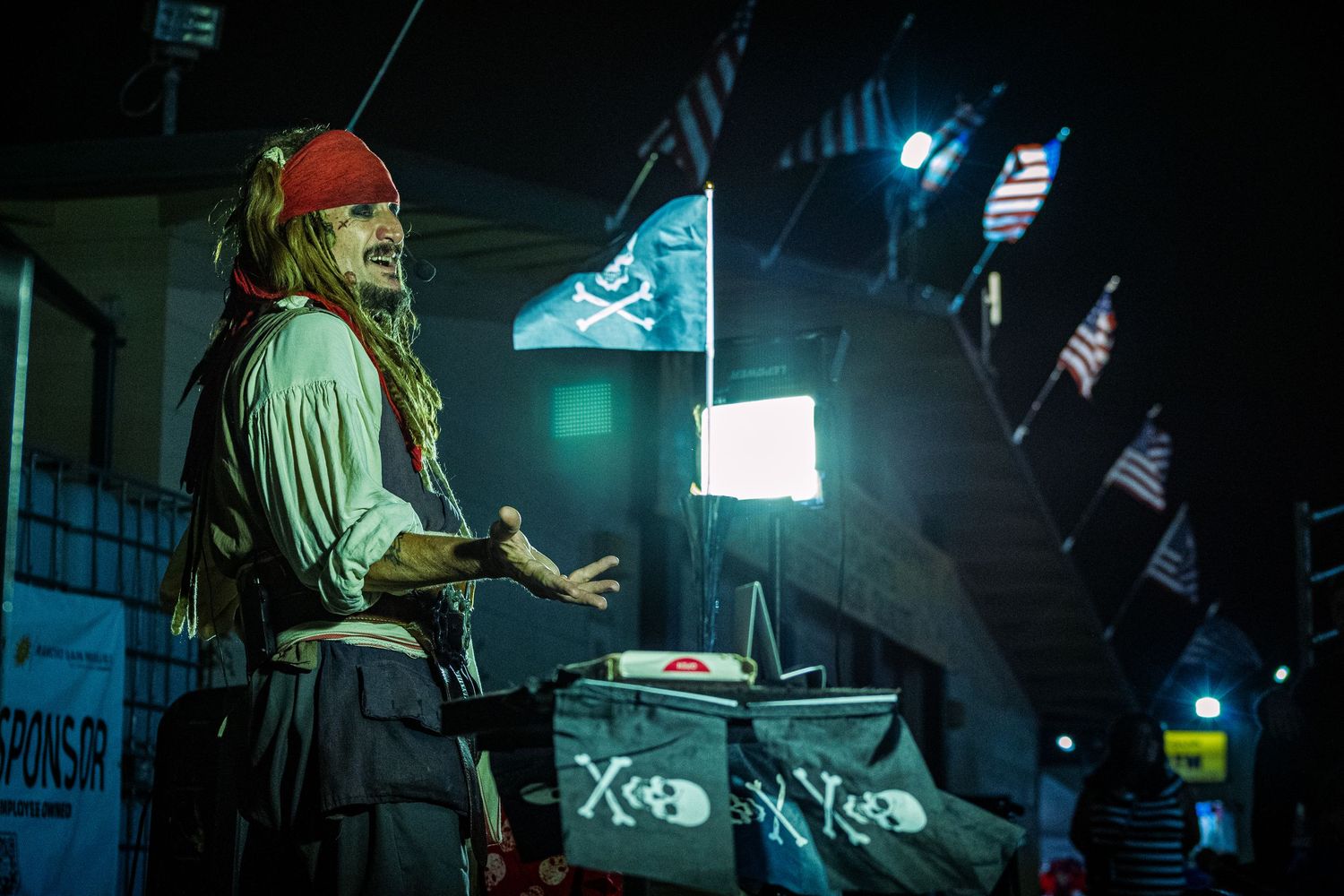 A pirate costume performer speaks at a nighttime outdoor event with skull flags and bright lighting.