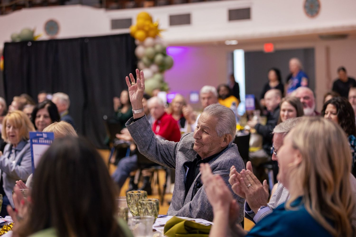 An elderly man raises his hand while surrounded by applauding guests at a festive indoor banquet event with balloon decorations.