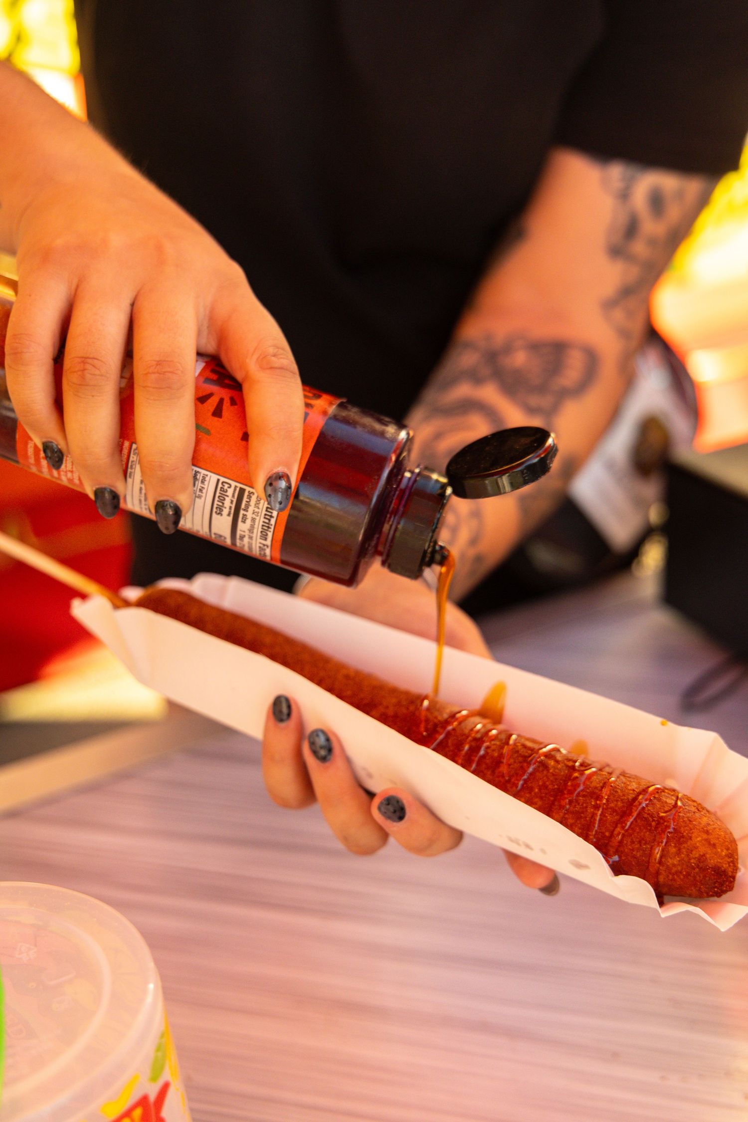 Tattooed hands pouring sauce over a corn dog in a paper tray at a carnival food stand.