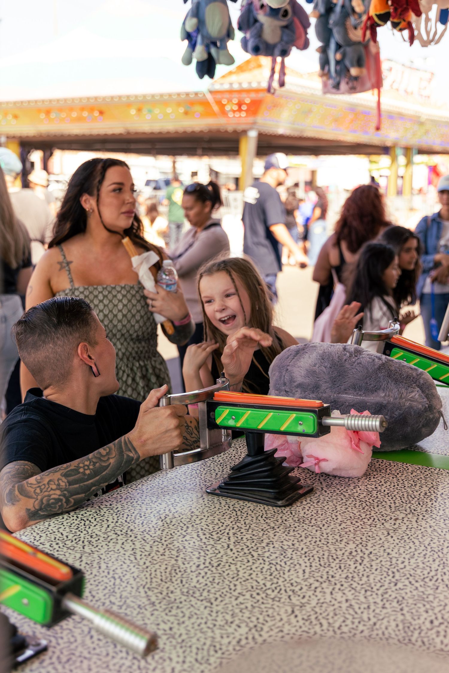 A tattooed man and excited young girl play a carnival game while a woman watches and smiles behind them.