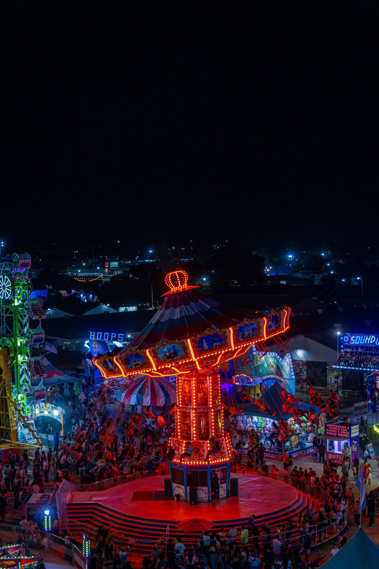 Aerial night view of a glowing red carnival ride surrounded by colorful fairground lights and crowds.