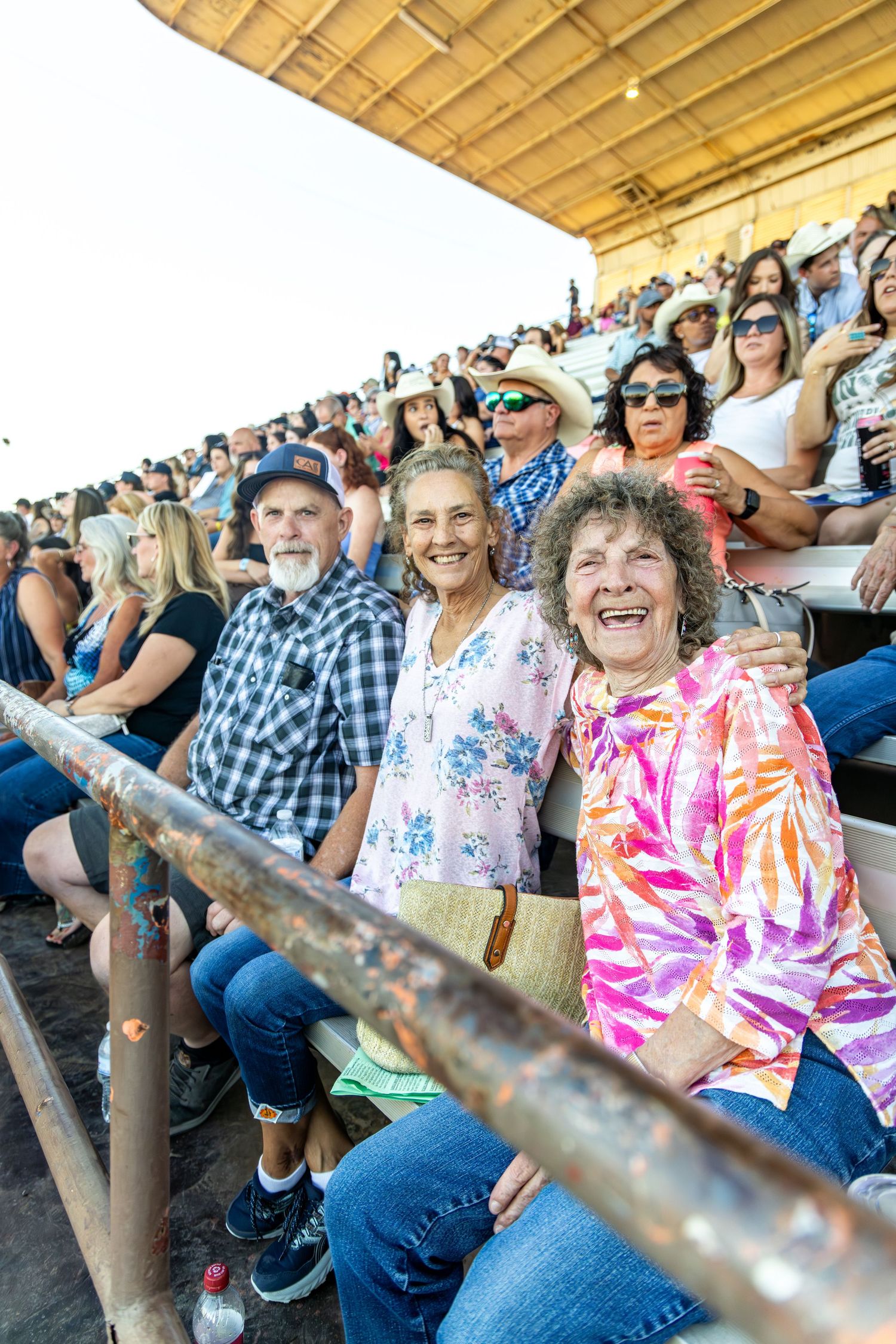 Three smiling seniors enjoy sitting together in packed bleachers at an outdoor fair event.