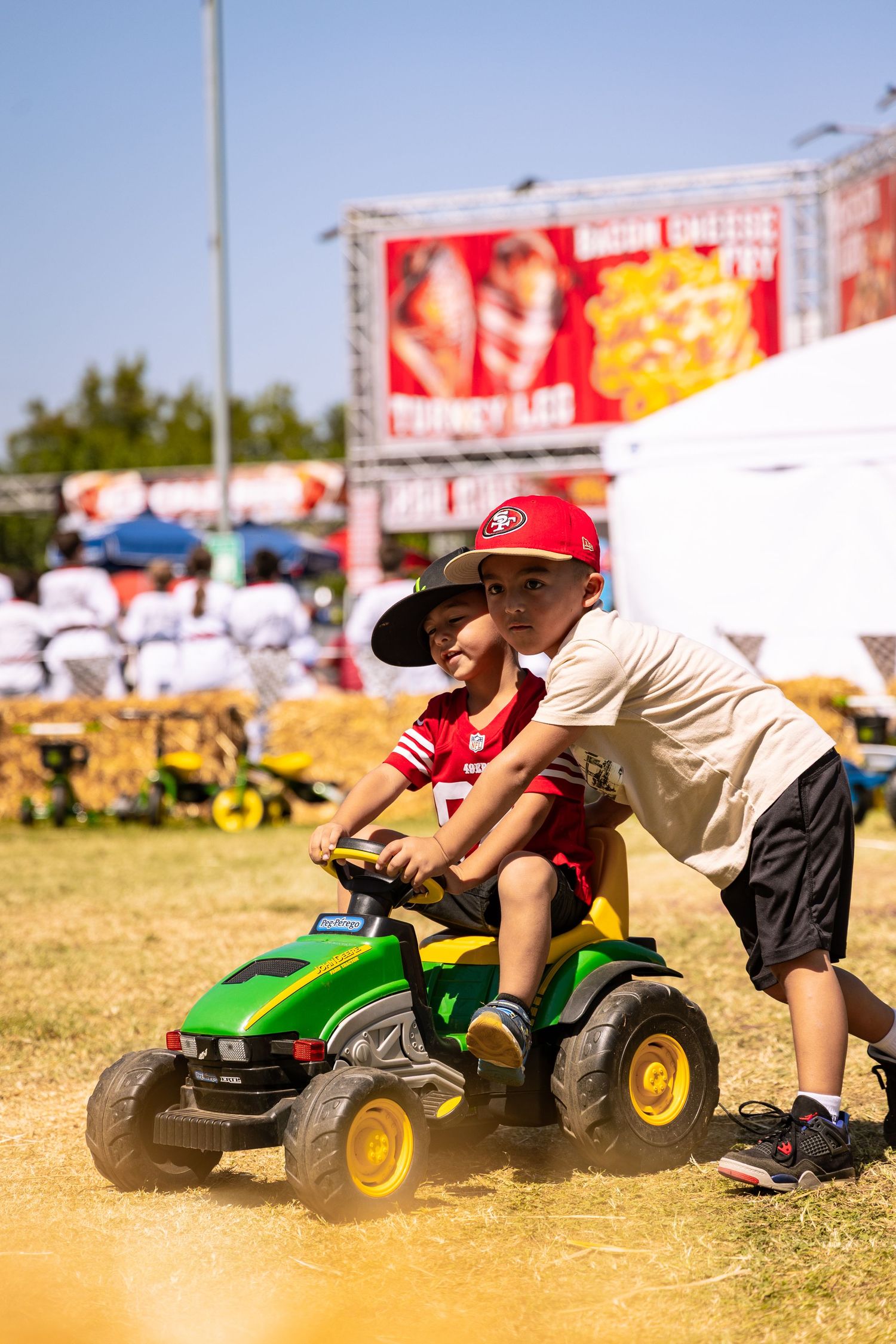 Two young boys play on a John Deere toy tractor at a sunny outdoor fair.