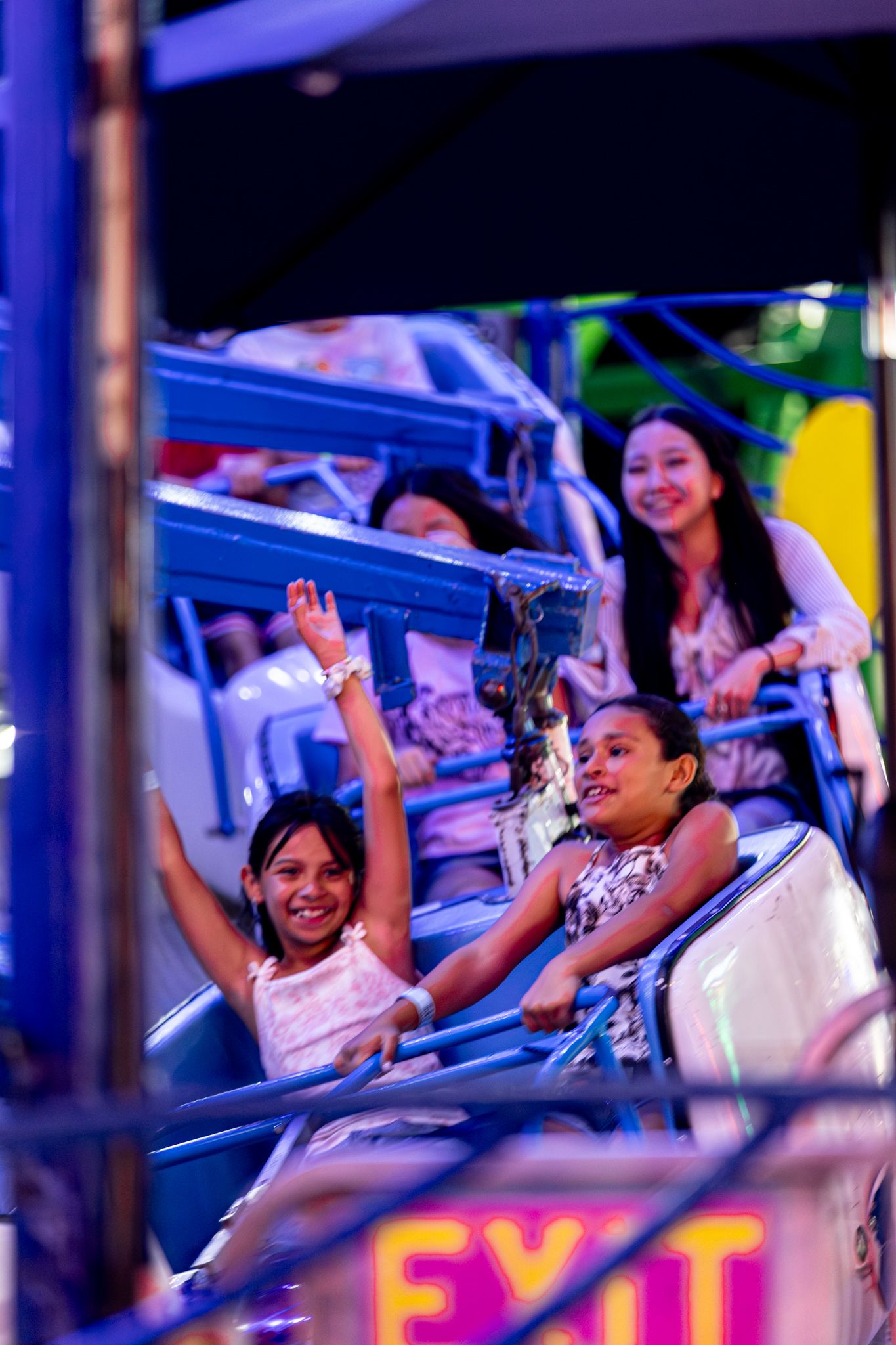 Two girls laugh and cheer on a carnival ride at a colorful amusement fair at night.