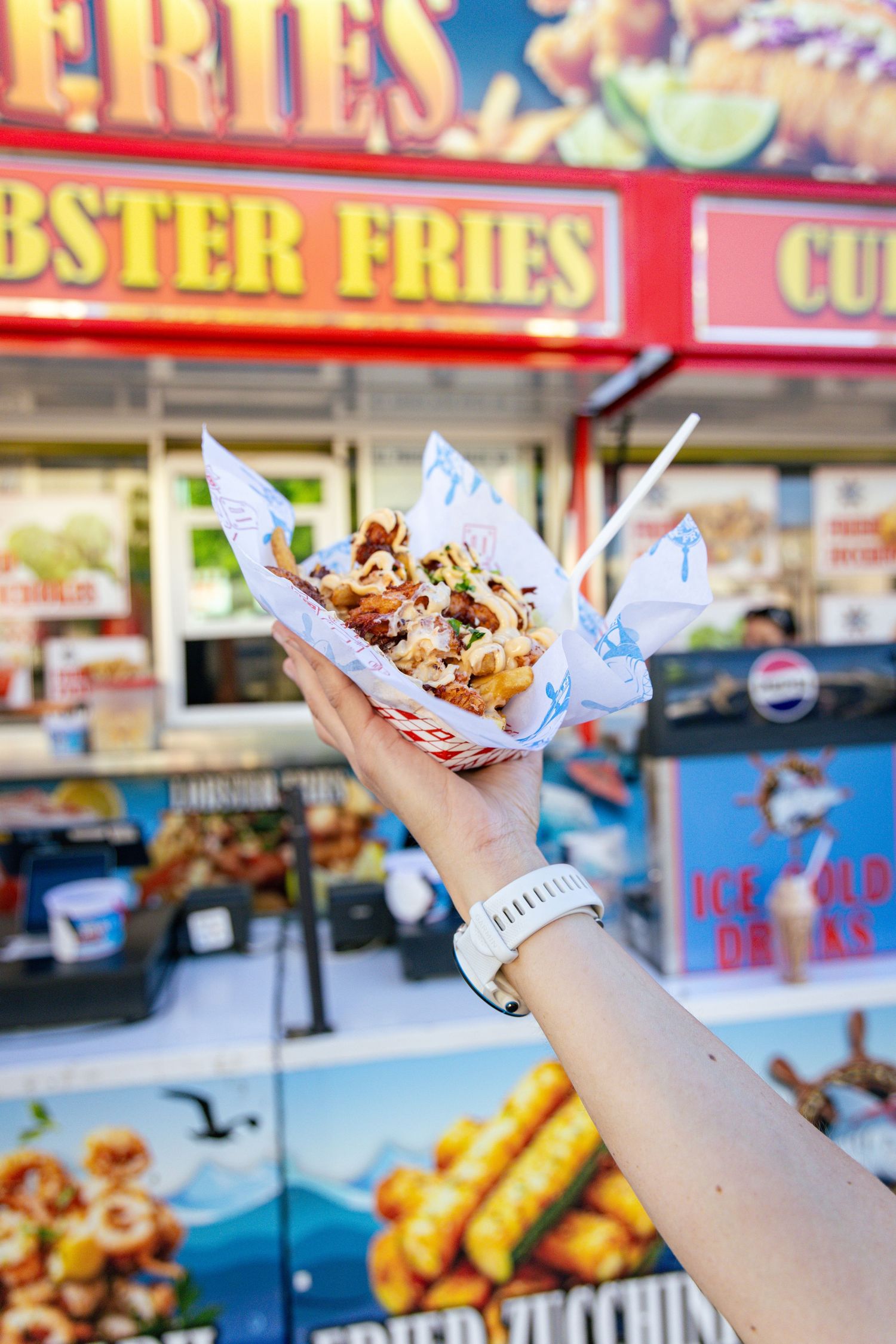 Hand holding a paper cone of loaded lobster fries with toppings in front of a colorful food stand.