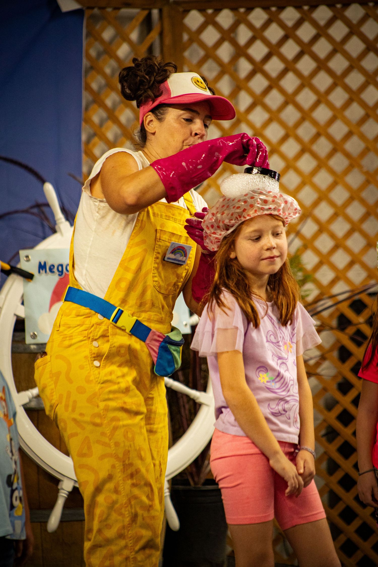 A performer in yellow overalls applies foam to a young girl's hair during a fun interactive children's show on stage.