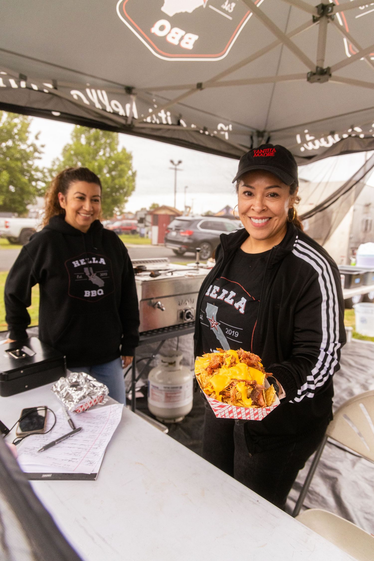 Two women in Hella BBQ branded black hoodies smile while serving nachos at an outdoor BBQ food stand.