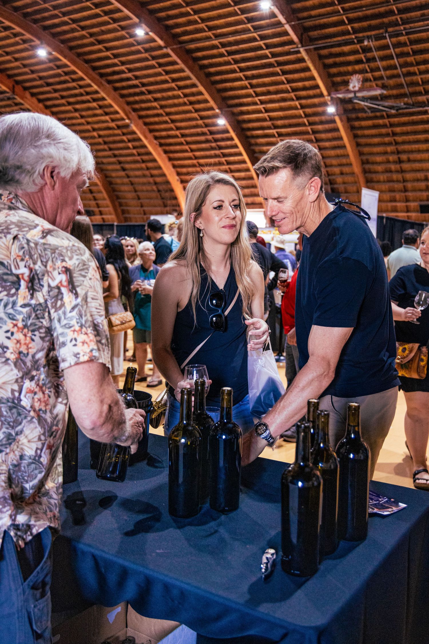 Guests sampling red wine at an indoor wine tasting event with bottles displayed on a draped table.