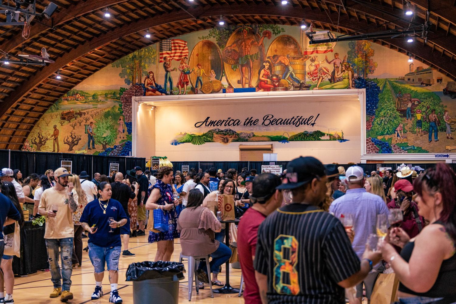 Crowds of people enjoying a wine tasting event inside a hall with an America the Beautiful mural.