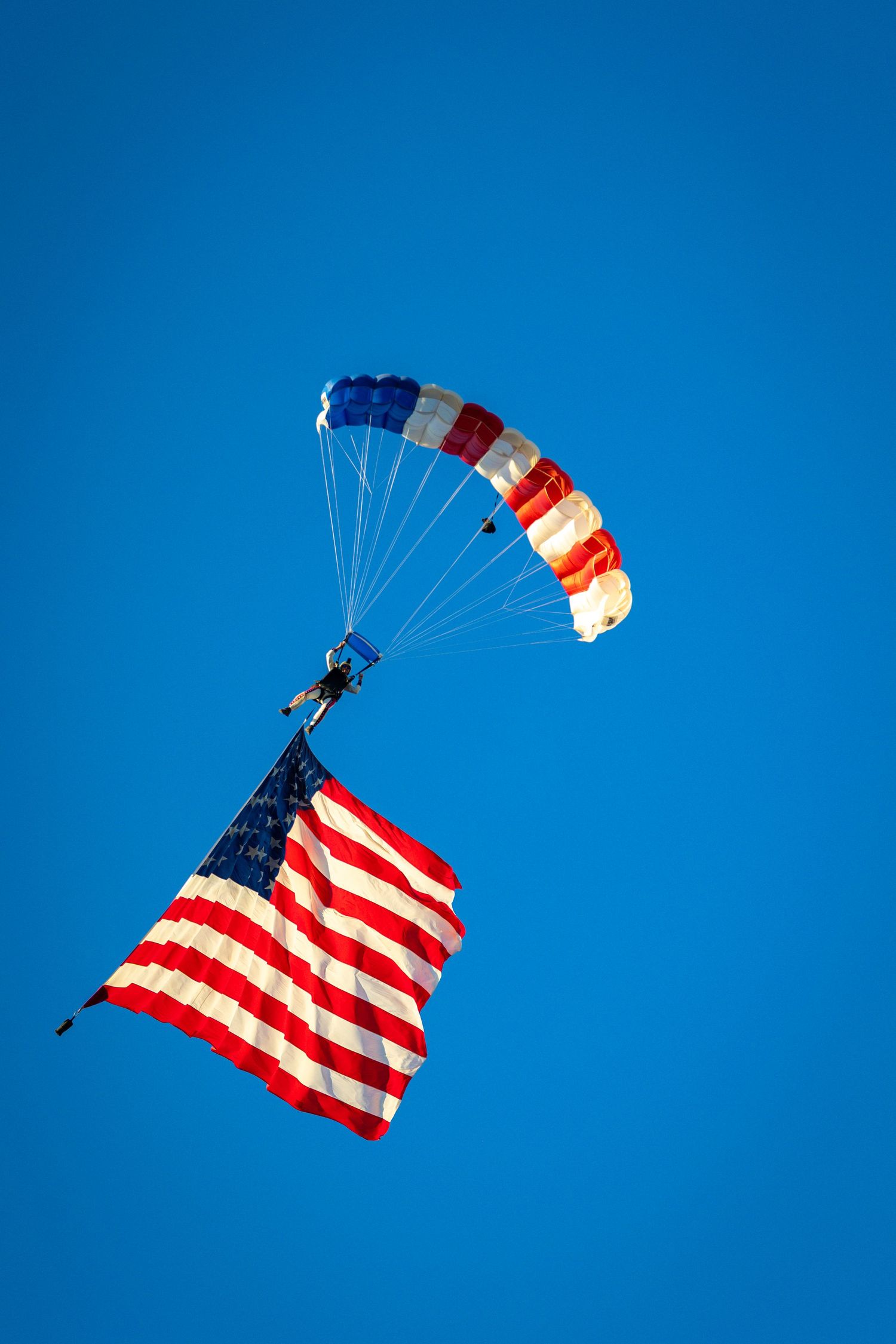 Skydiver carrying a large American flag descends with a colorful parachute against a clear blue sky.