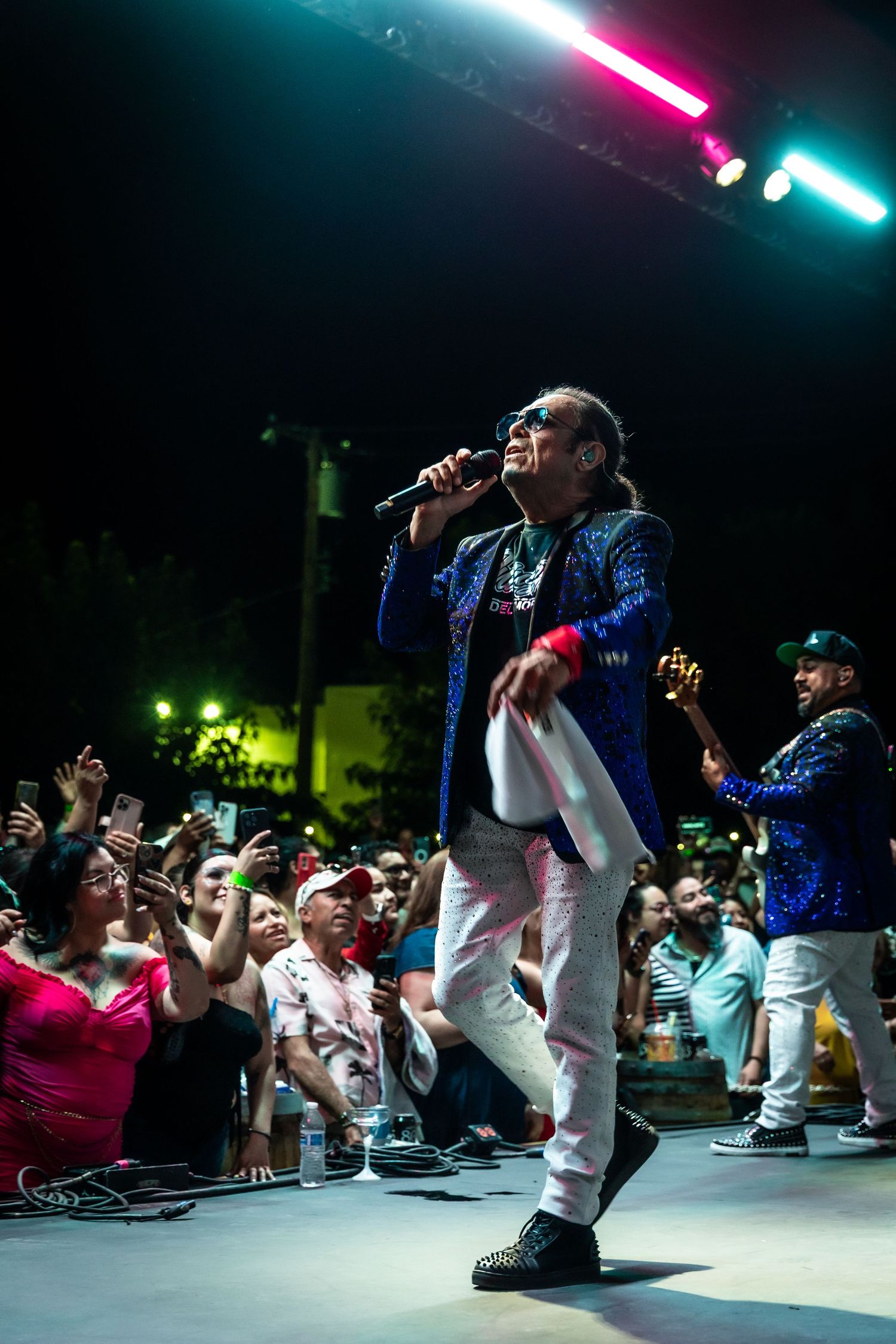 Energetic male singer in a blue sequin jacket performs on stage before a large outdoor night crowd.
