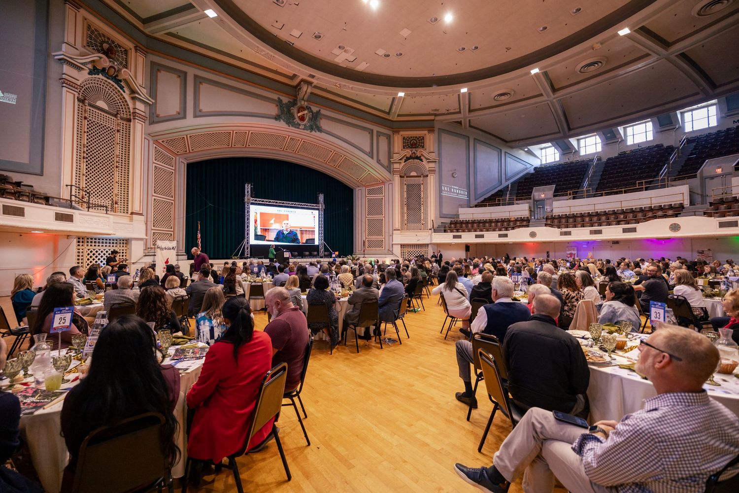 Large formal banquet event in a grand hall with hundreds of guests seated at round tables facing a presentation screen.