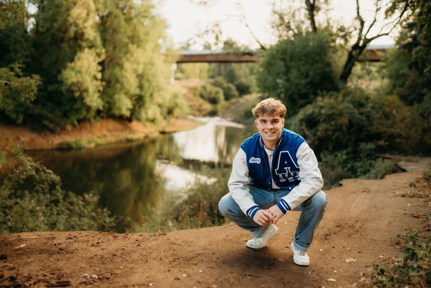 Senior boy crouching by a river with natural, relaxed expression.
