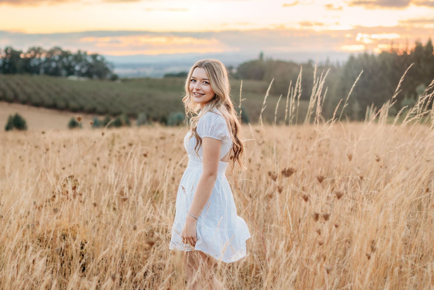 Senior girl standing in tall grass wearing a white dress at sunset