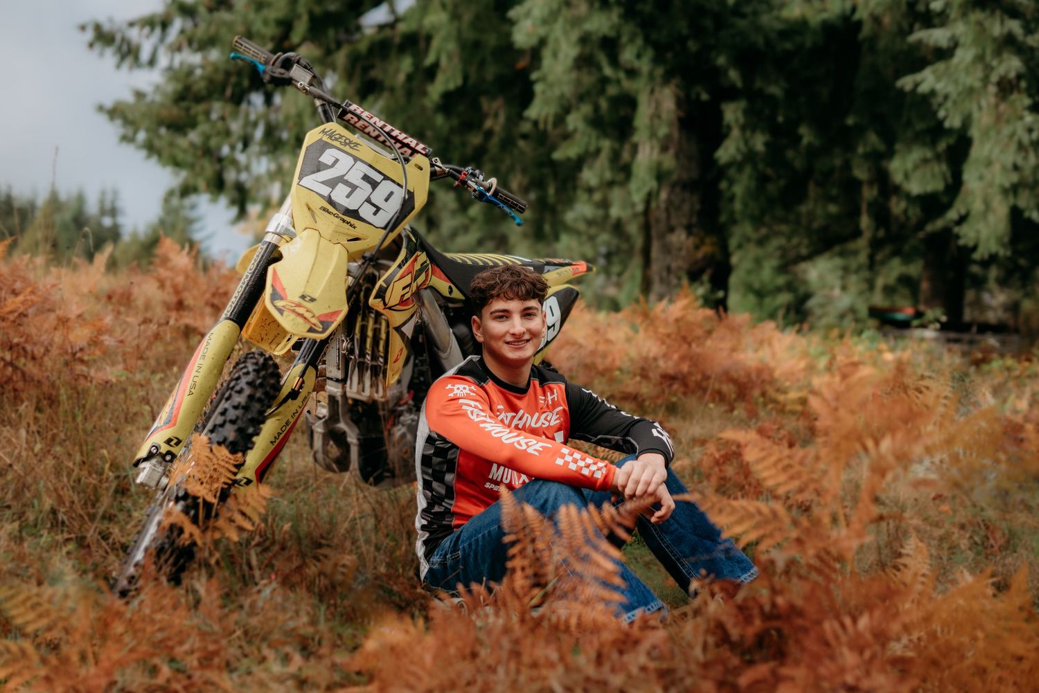 Senior boy sitting and smiling next to his dirt bike in a field
