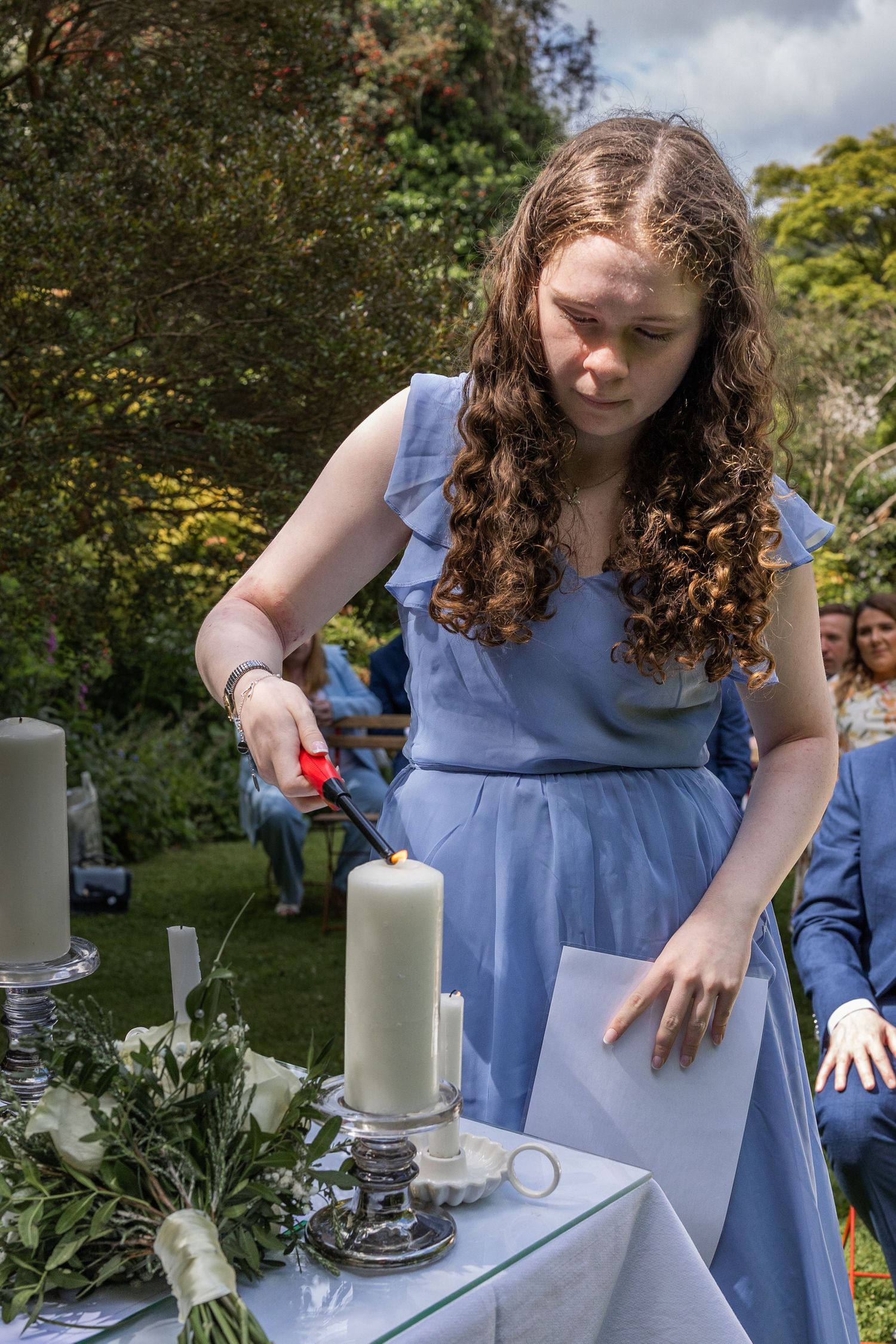 Young woman in blue dress lighting a candle at an outdoor wedding ceremony with floral decorations.
